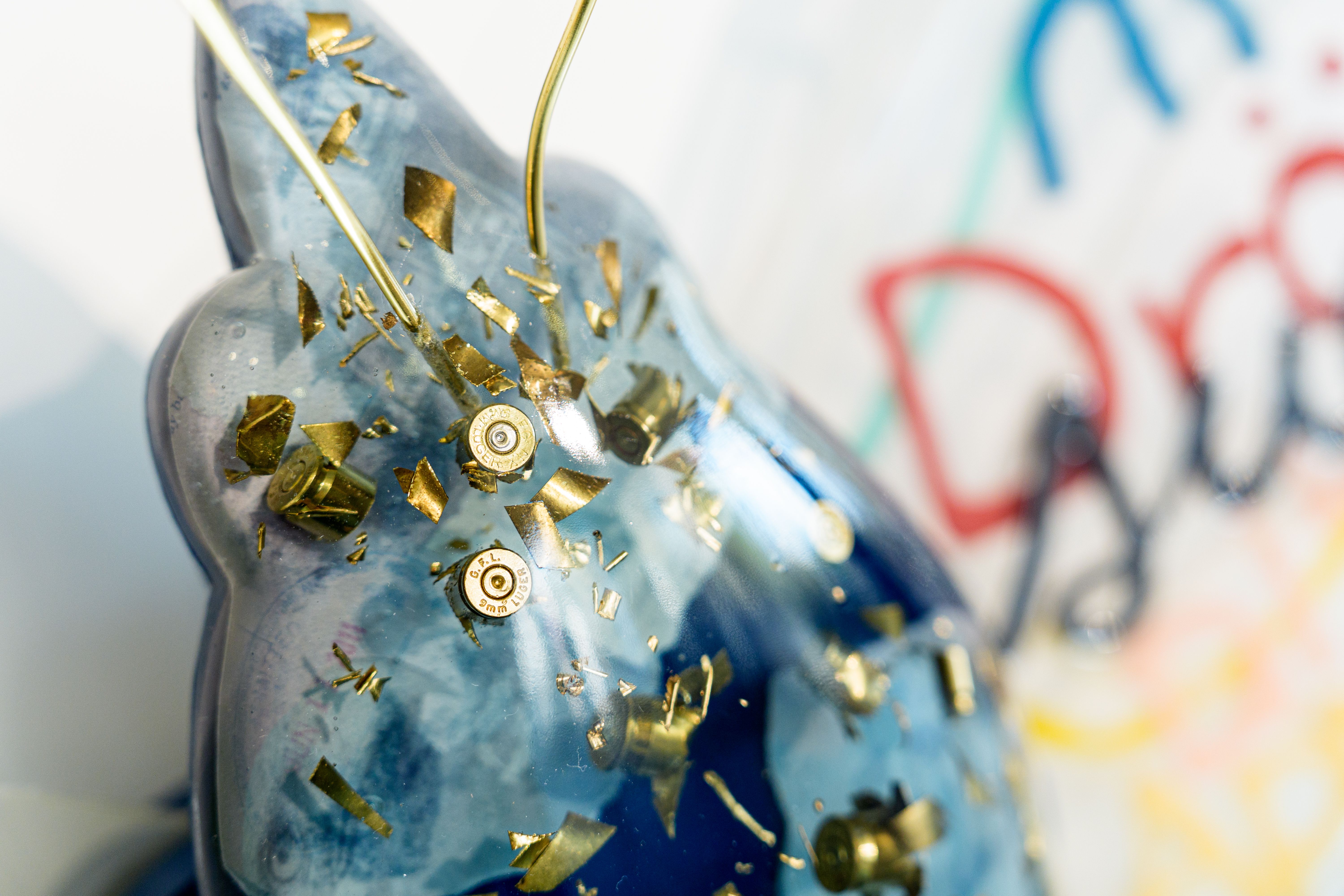 Close-up of a blue sculpture with embedded gold-colored bullet casings and fragments, against a blurred background with red, blue, and green graffiti text.