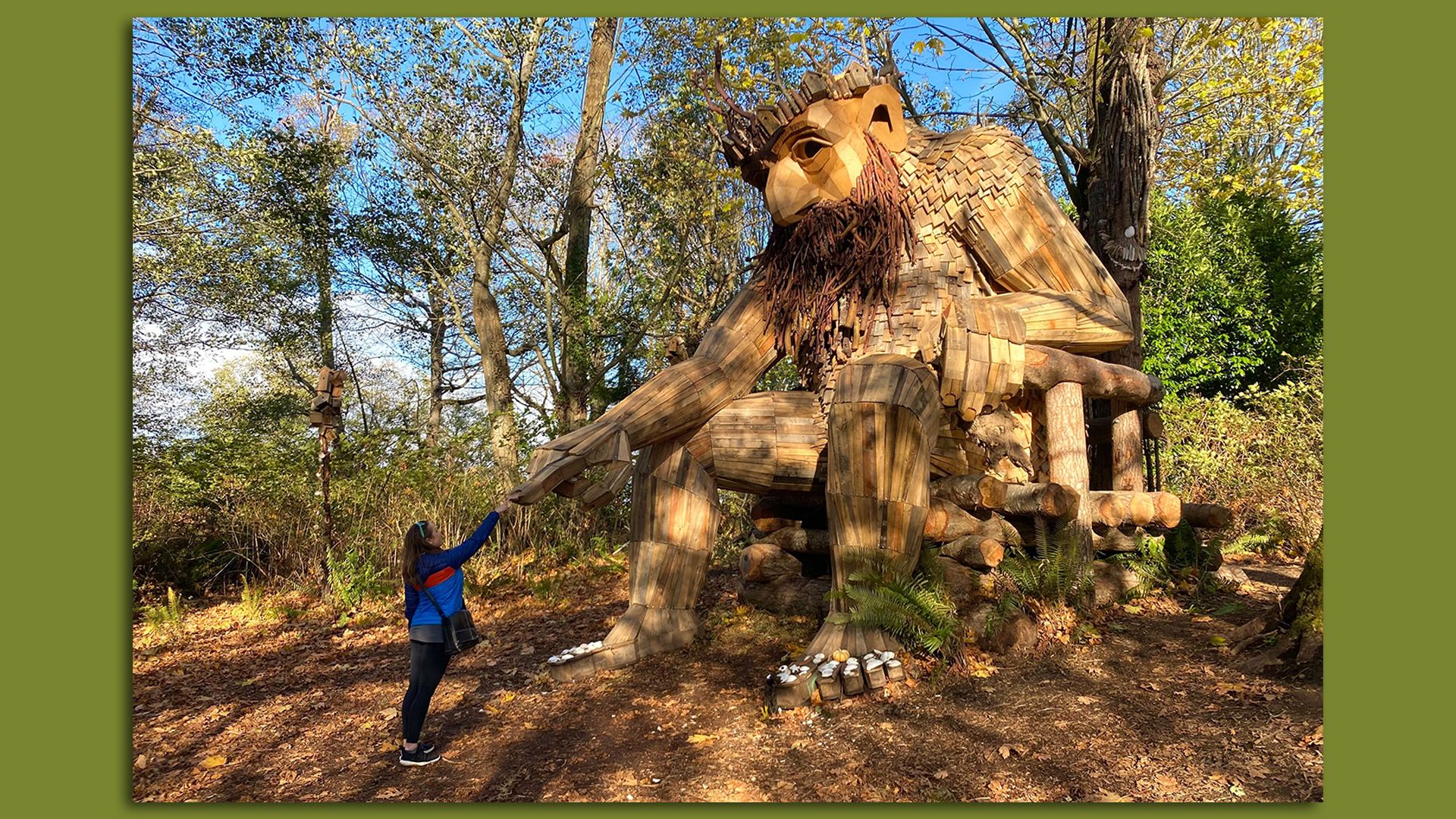 A towering wooden troll sits on a throne surrounded by trees as a woman reaches out to touch its hand.