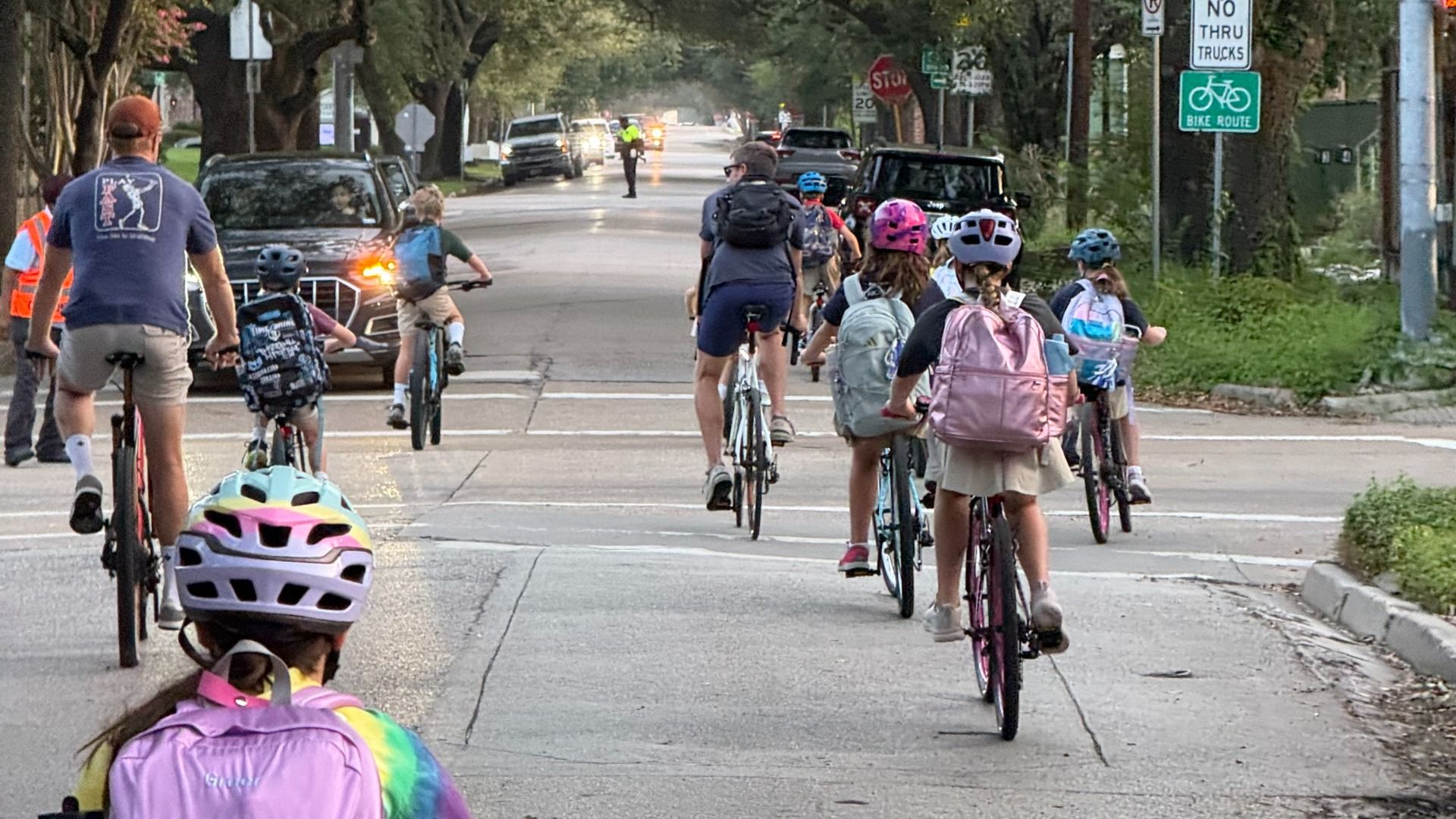 Group of children wearing helmets and backpacks ride bicycles down a tree-lined street as cars wait; a stop sign and a bike-route sign are visible ahead.