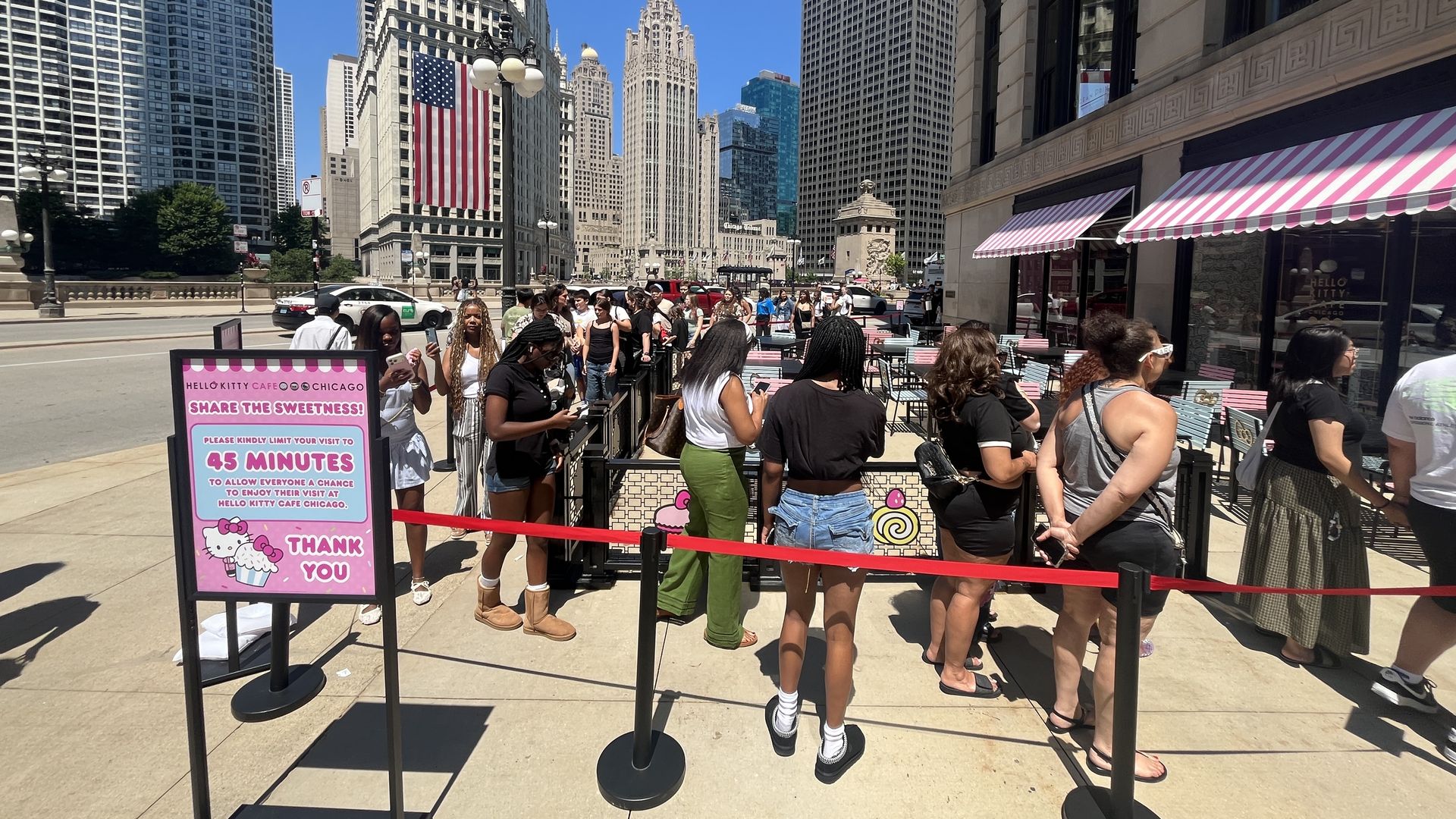 Busy city street with tall skyscrapers, a clock tower, and a large American flag on a historic building. A crowd lines up beside pink-and-white striped cafe awnings; Hello Kitty sign in foreground.