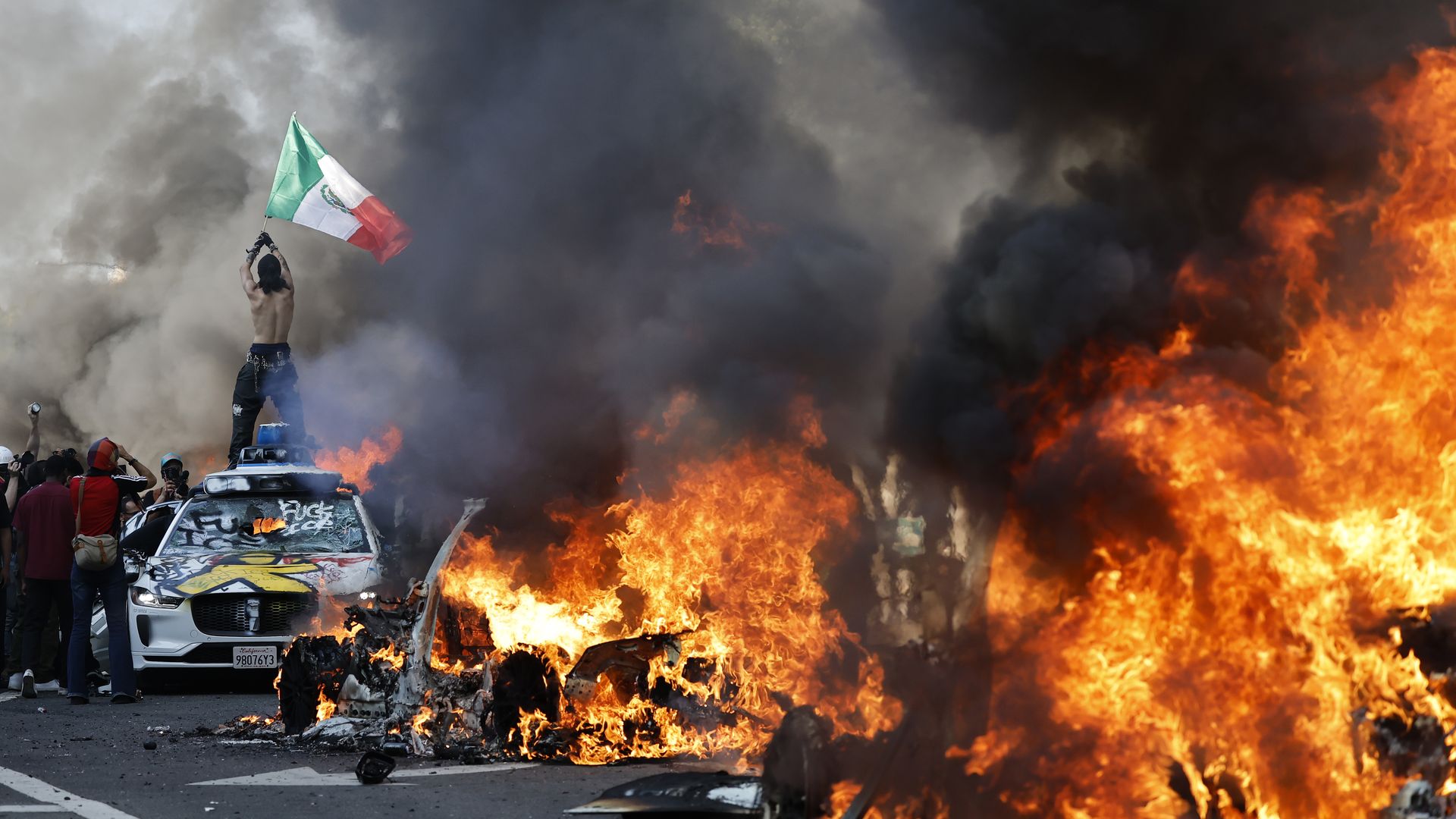 A protester stands on top of a car holding a Mexican flag as burning vehicles line the street.