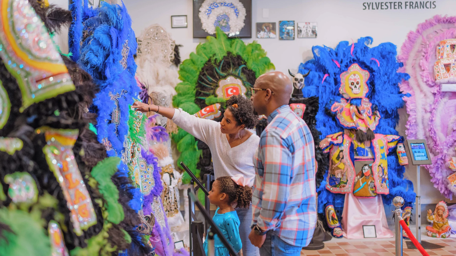 A family of three—a woman, man, and young girl—observe colorful, feathered Mardi Gras Indian costumes displayed in a museum setting with vibrant blue, green, purple, and pink hues.