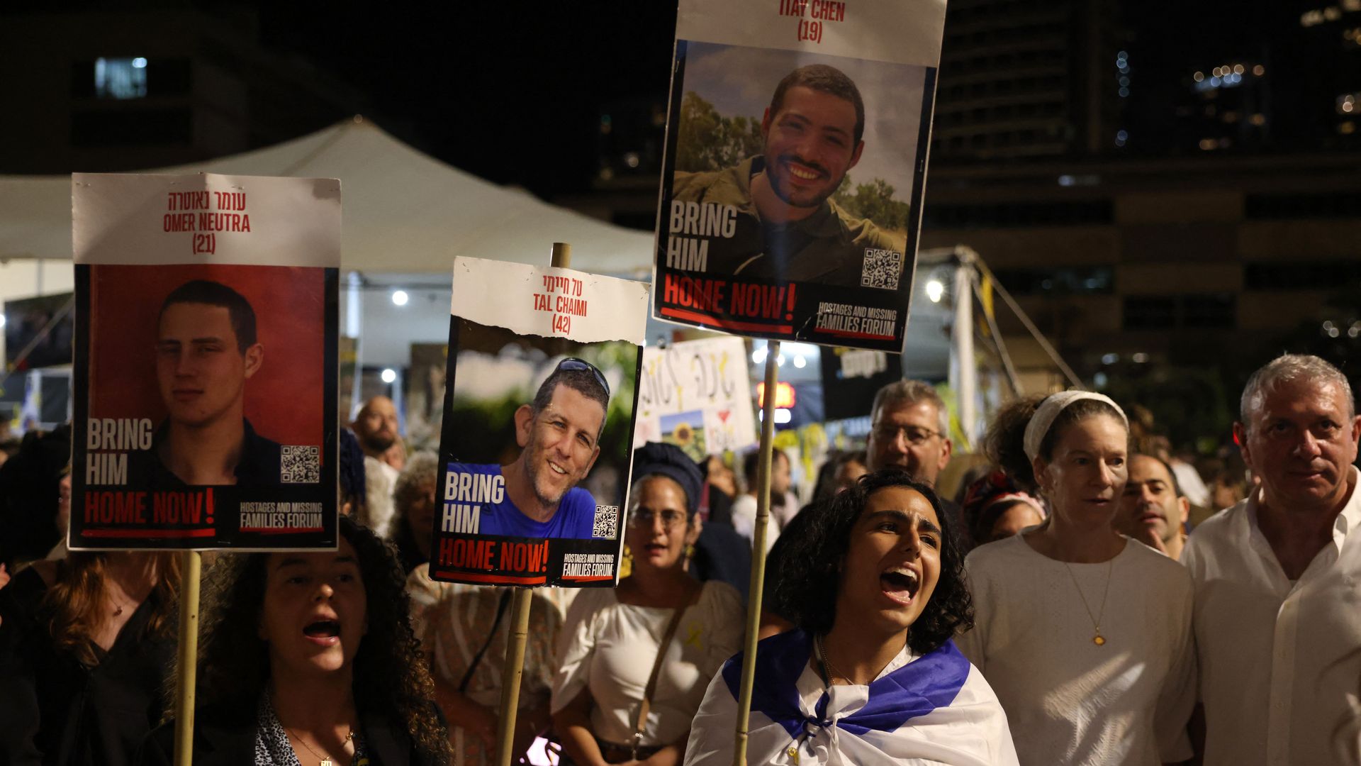 Protesters, wearing white and black, hold signs with men's faces on them with the words "BRING HIM HOME NOW!"