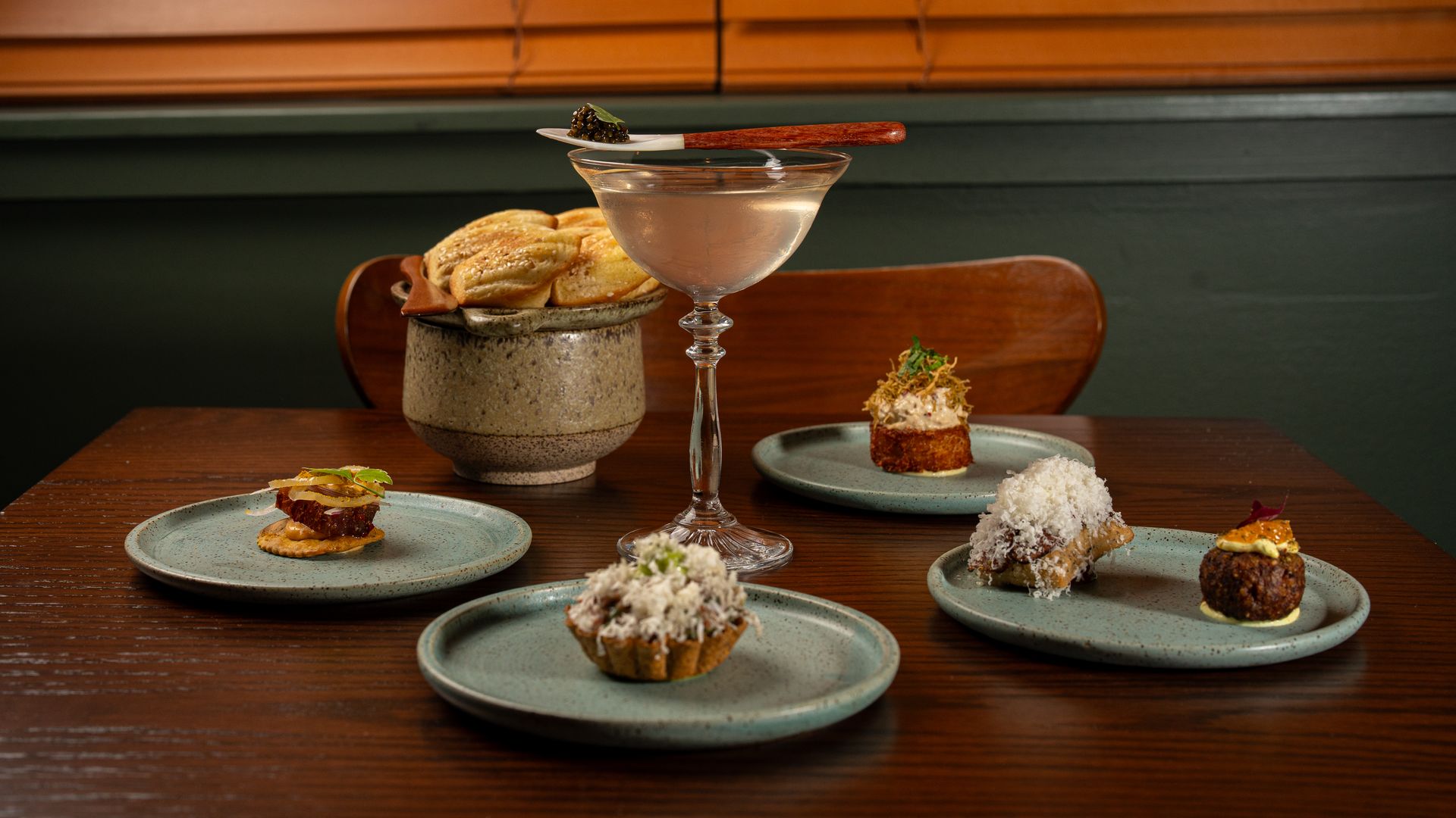 A wooden table with five plates holding assorted gourmet appetizers and a clear cocktail in a glass with a small knife resting on top, set against a dark green wall and closed orange blinds.