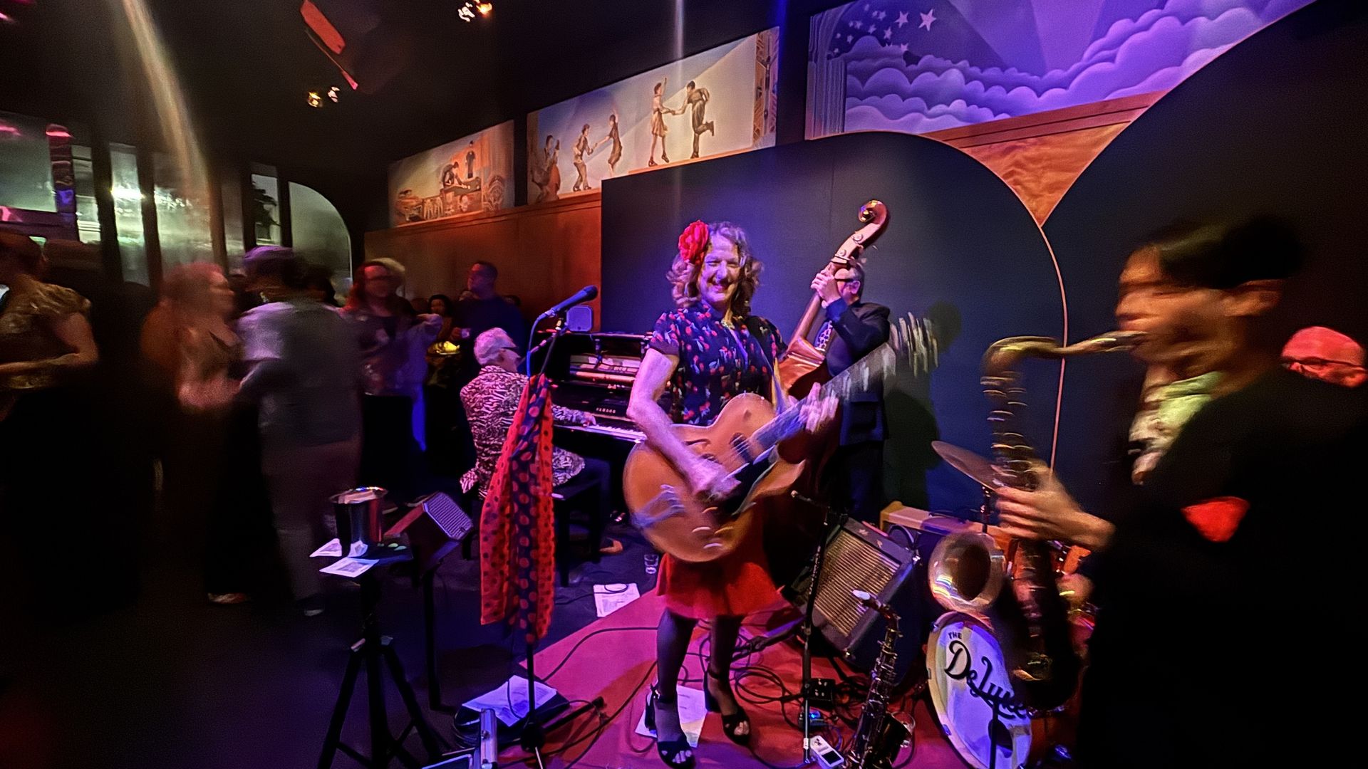 Live band on a small stage in a dim, colorful venue; smiling guitarist with a red scarf, lead singer at the mic, bass player nearby; crowd dancing, purple wall mural and warm stage lights.