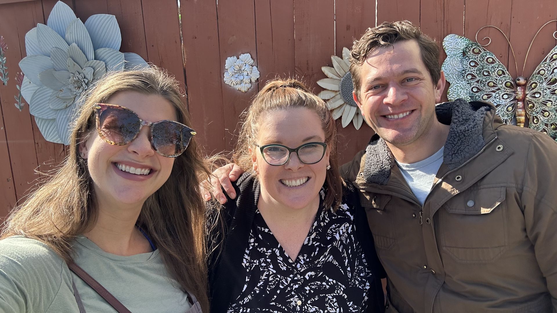 Photo shows Chelsea Brasted, Carlie Kollath Wells and Alex Thompson smiling in front of an art-covered fence.
