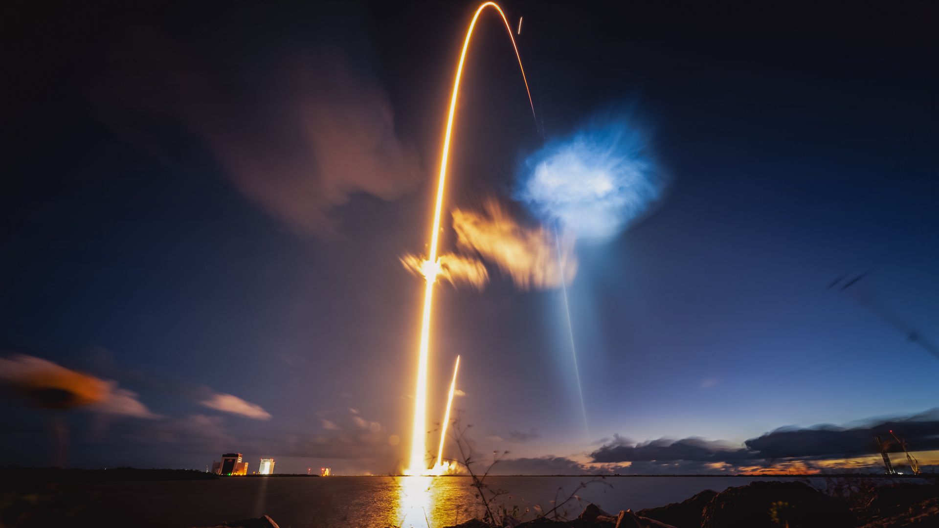 Long-exposure night launch over water: an orange rocket plume forms a bright arc into the dark sky, with blue smoke to the right; shoreline rocks and distant buildings glow below.