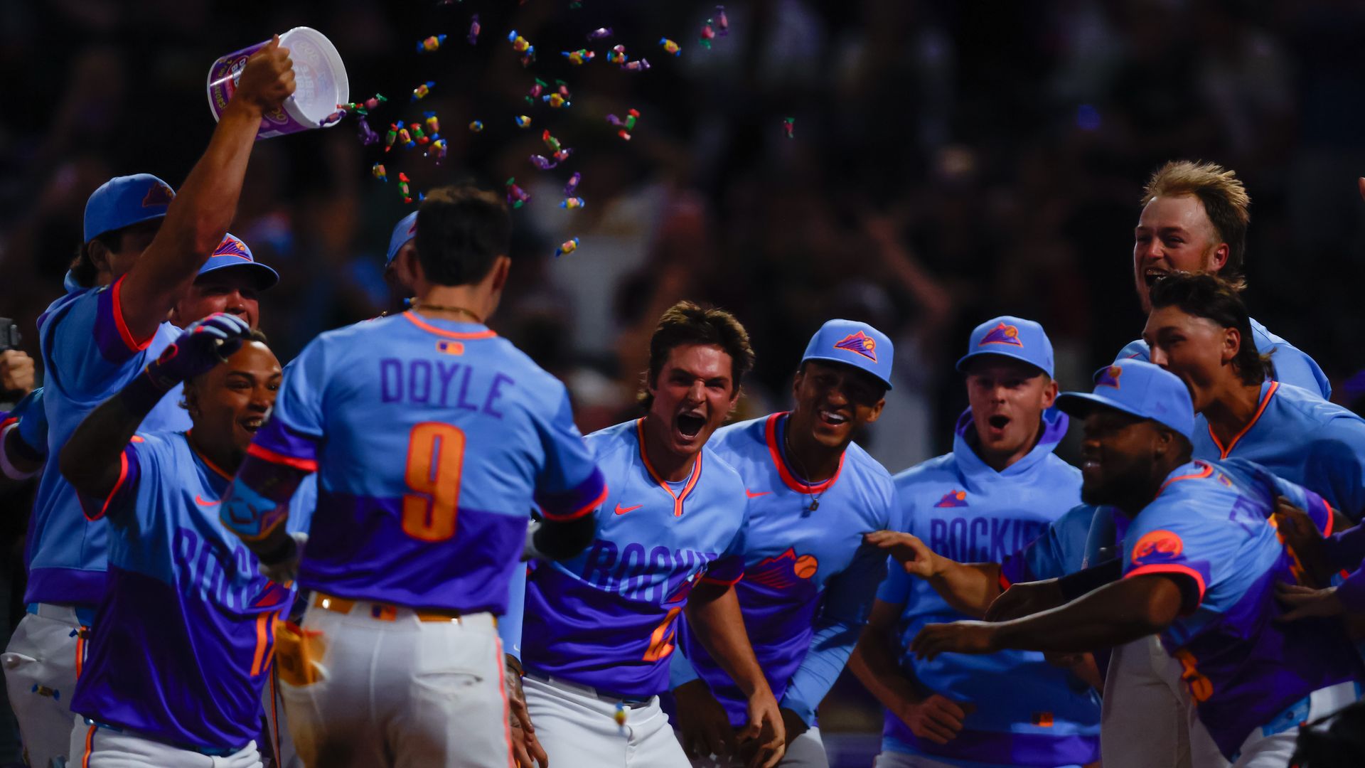 Colorado Rockies baseball players in blue and purple uniforms celebrating energetically, one player spraying colorful confetti over teammate with number 9, Doyle, at night.