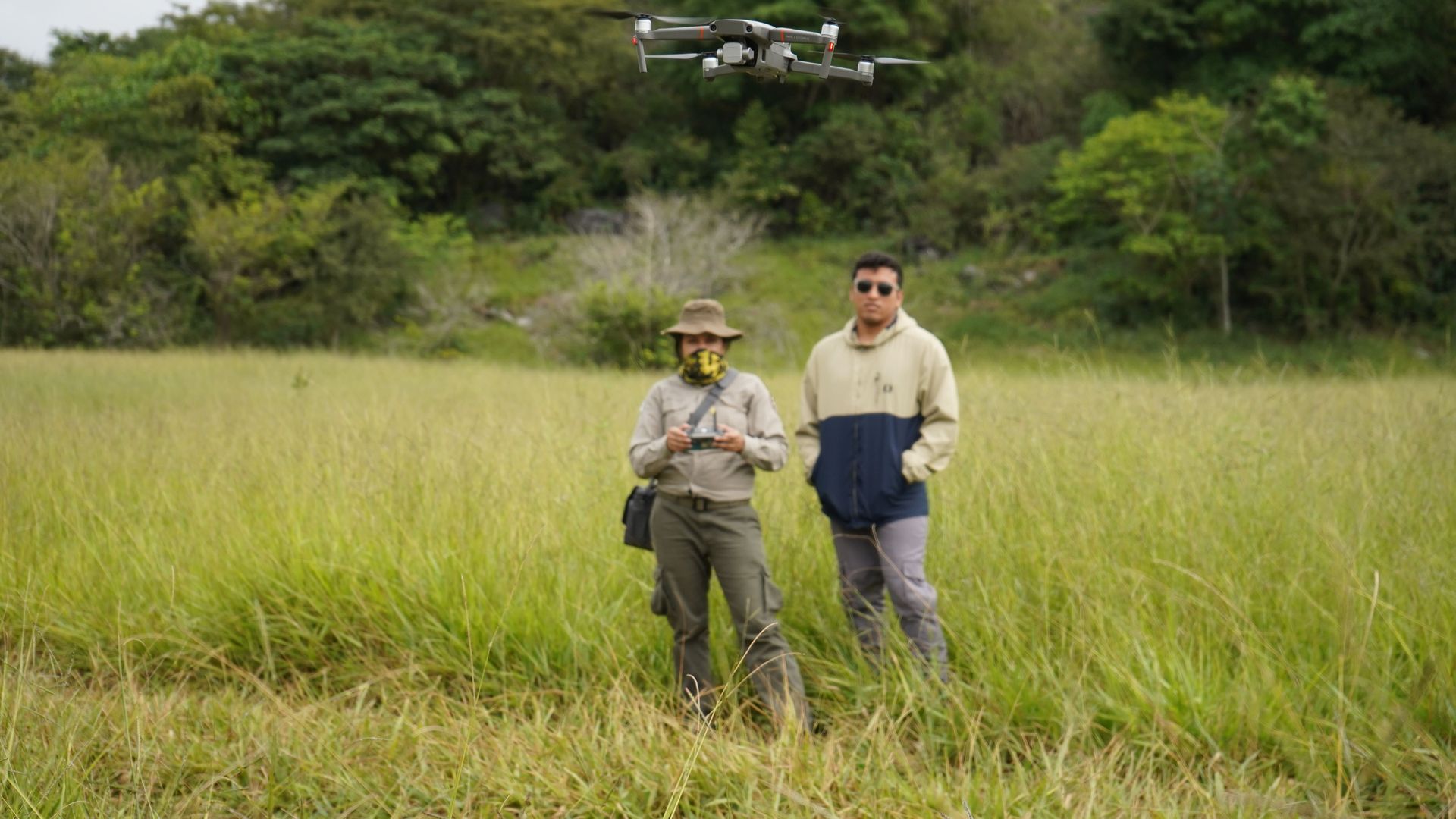 A person pilots a low-flying drone while another person stands on in the Amazon rainforest