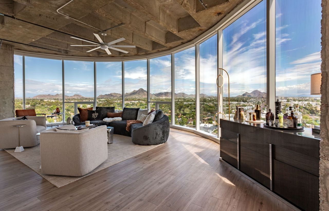 Living room with curved floor-to-ceiling windows showing city and mountains. Beige and dark gray sofas around a light rug; a bar with bottles on the right; exposed concrete ceiling and a white ceiling fan.