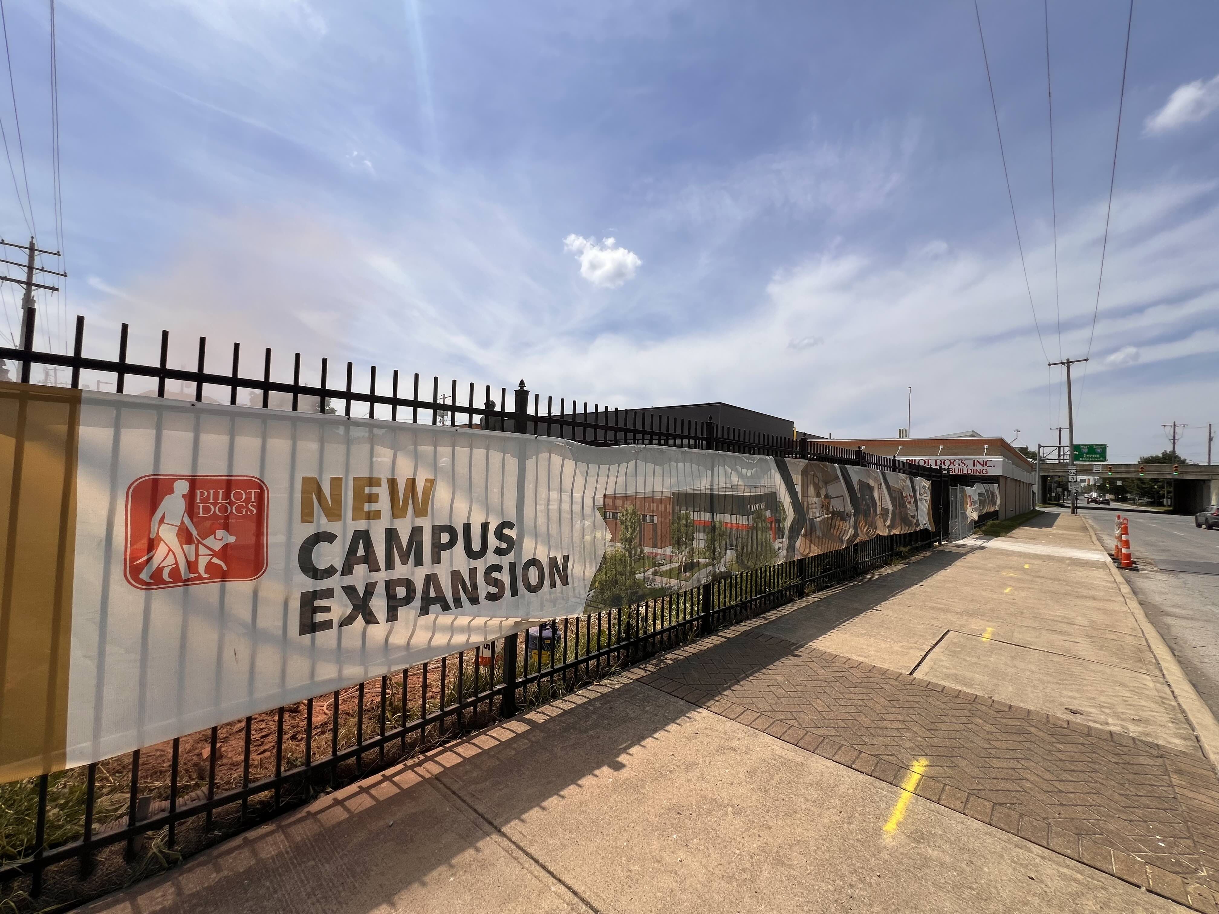 Fence banner outside Pilot Dogs building announces "New Campus Expansion" with logo of person walking a dog, under a blue sky with some clouds along a city sidewalk.