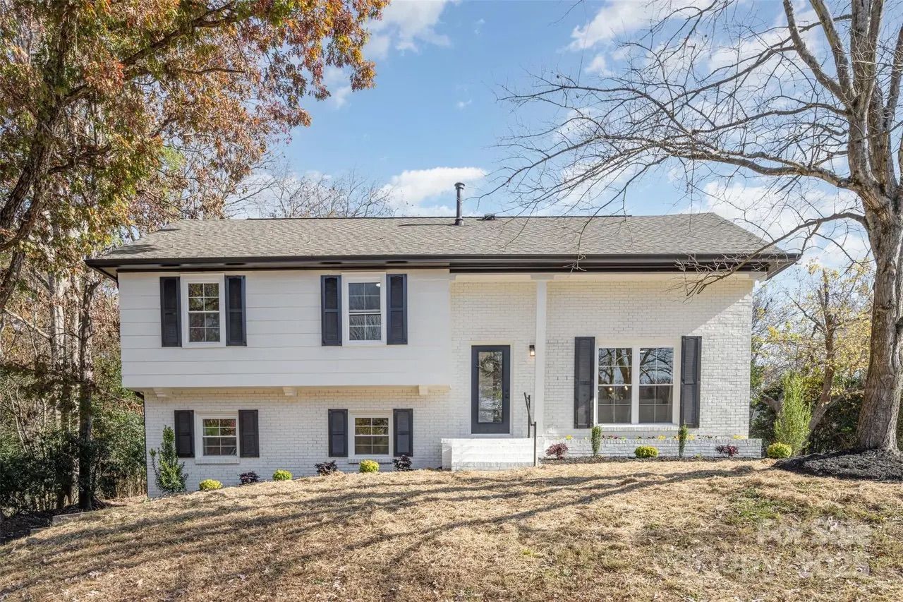 White two-story house with black shutters and front door, surrounded by trees with autumn leaves and a dry grass lawn under a blue sky with clouds.