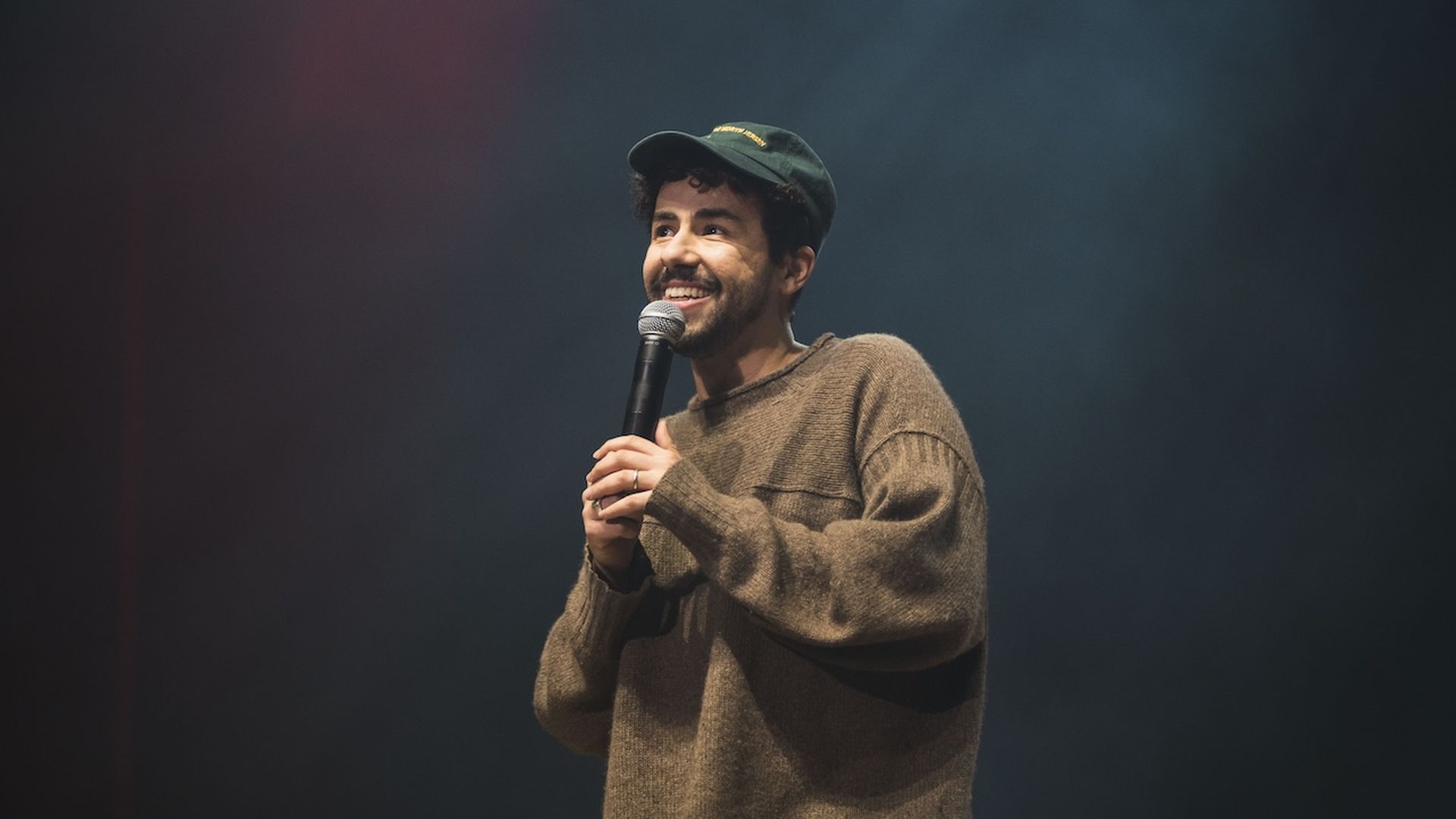 A photo of comedian Ramy Youssef holding a microphone with a black stage background behind him.