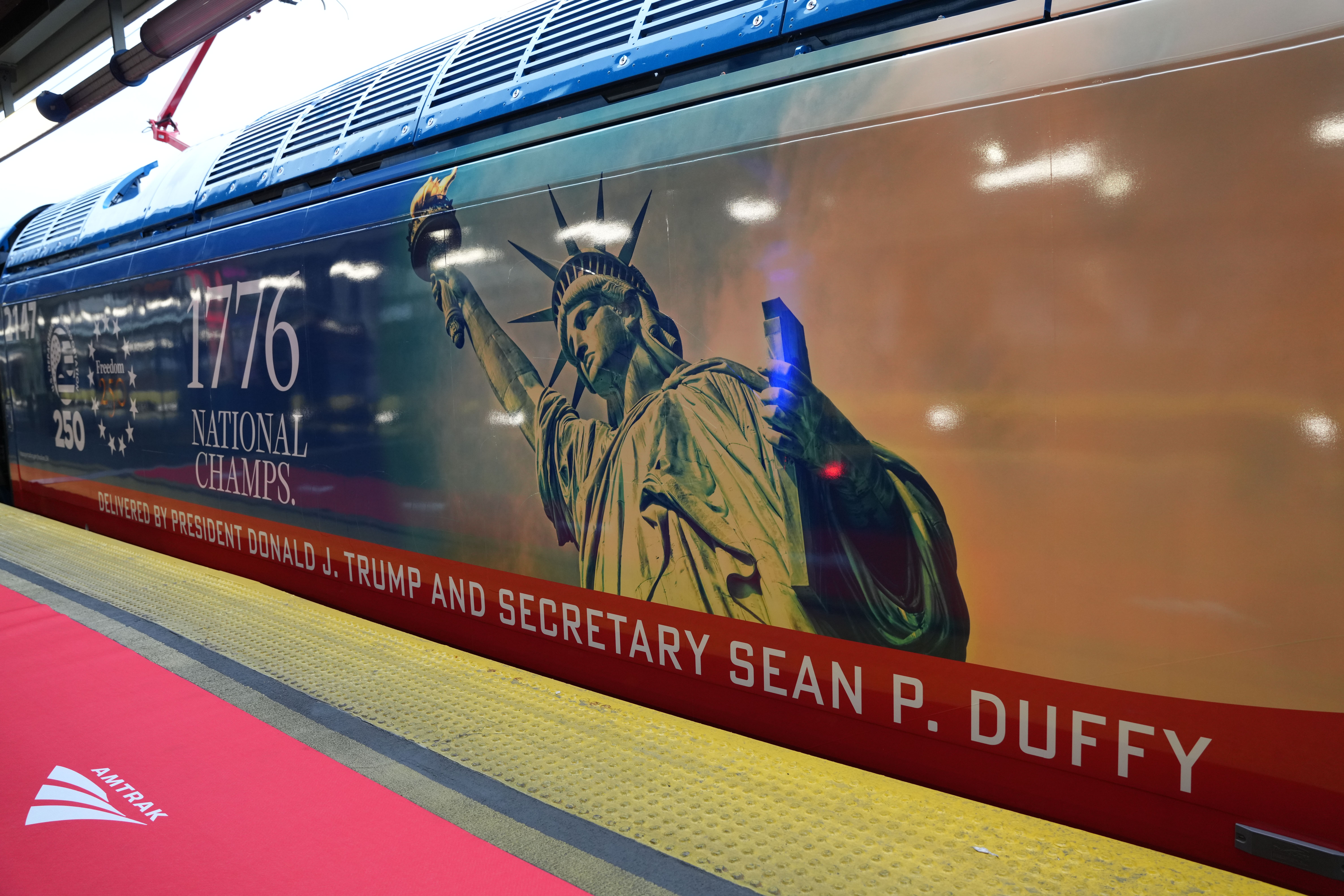 Side of an Amtrak train wrap featuring a Statue of Liberty graphic, large 1776 and "National Champs" text, blue roof vents, a red promotional stripe, and a yellow textured platform edge with the Amtrak logo.