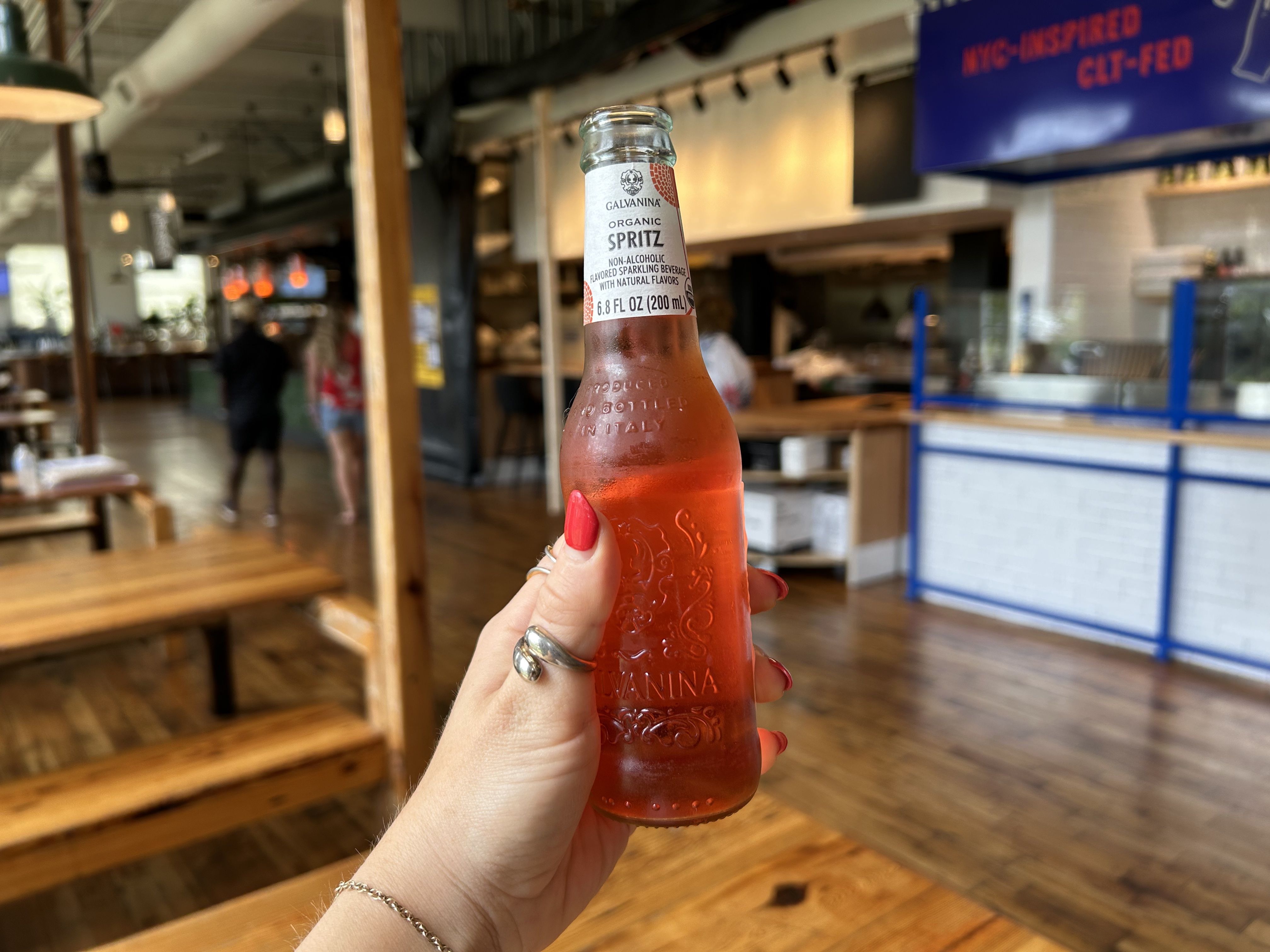 Hand with red nail polish and silver rings holding a bottle of Galvanina Organic Spritz, a non-alcoholic sparkling beverage, inside a casual restaurant with wooden tables and people in background.