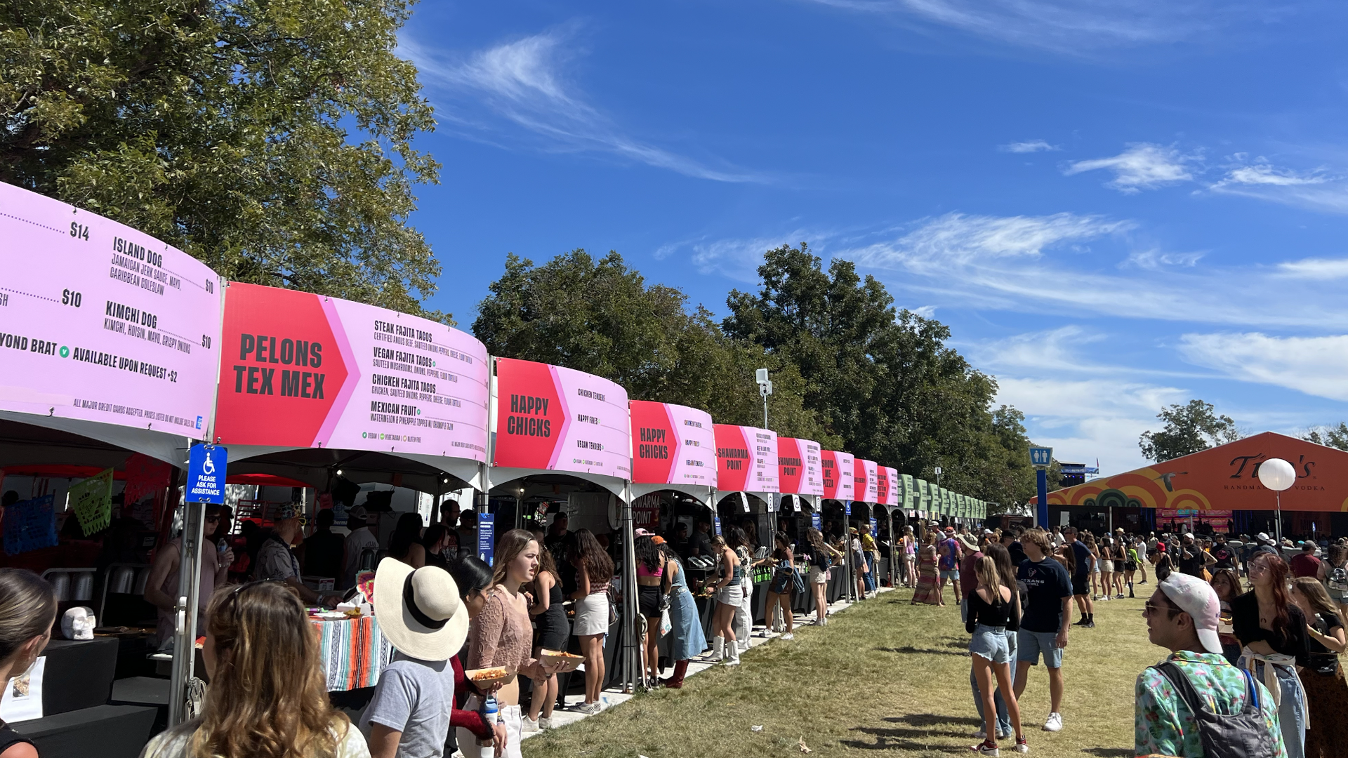 Outdoor food festival with people in line at colorful pink and green food tents under a bright blue sky with wispy clouds and large trees in the background.