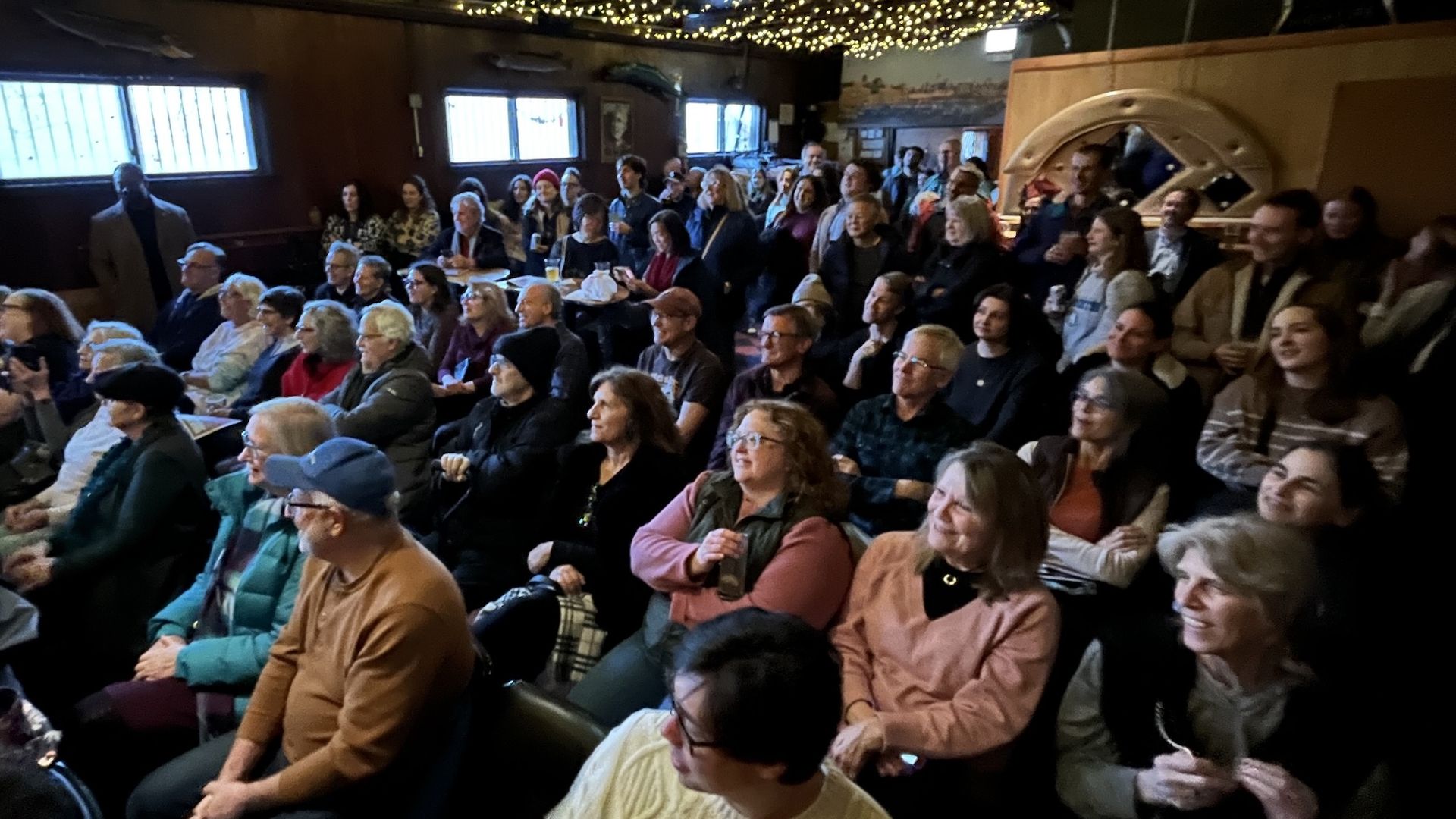Crowd of adults seated and standing in a cozy room with warm string lights and a disco ball overhead; wooden walls, large windows, and a circular decor piece at the back right.