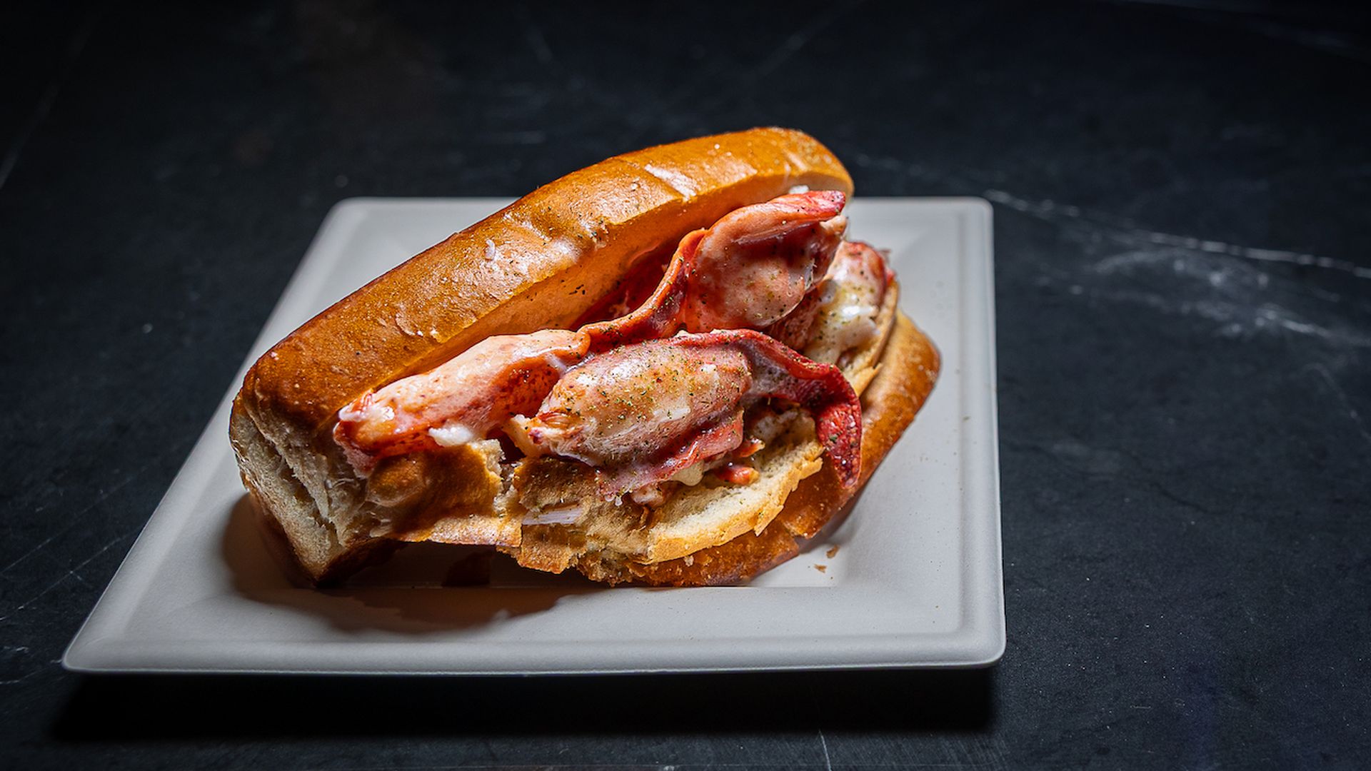 A lobster roll with a toasted bun overflowing with pink lobster meat, mayo and herbs, served on a white square plate on a dark, textured surface.
