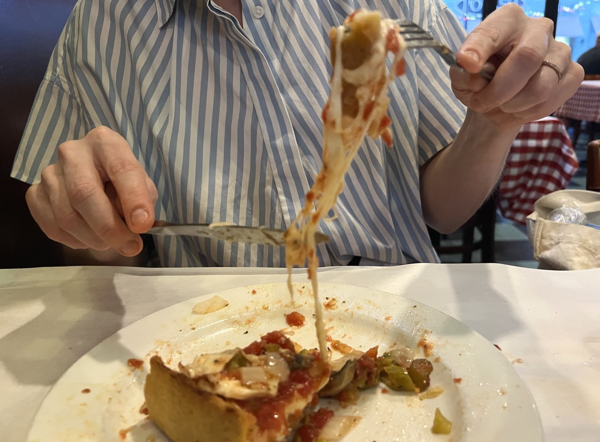 A person in a blue-and-white striped shirt uses a fork to lift a long, cheesy pizza slice. Toppings resemble peppers and tomatoes; a checkered tablecloth is visible in the background.