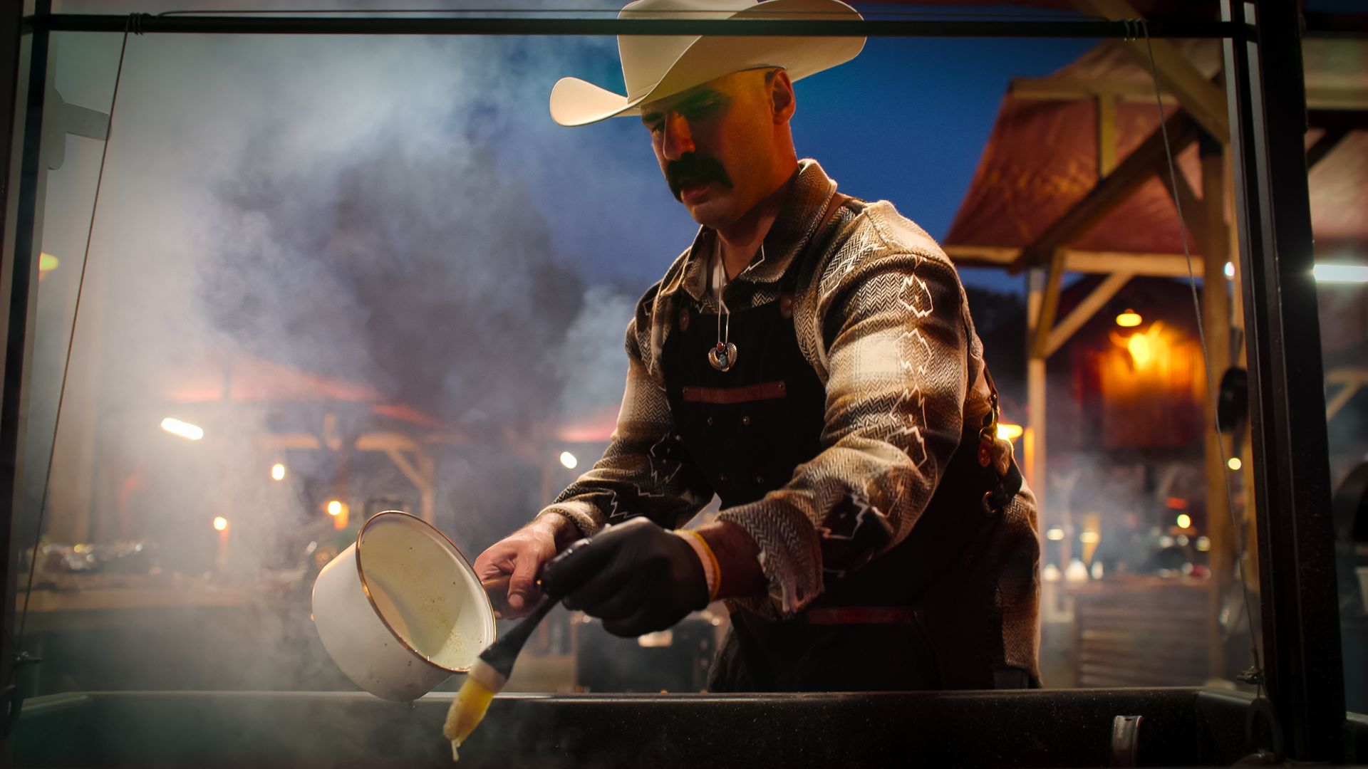 A photo of a person in a cowboy hat standing over a grill with a white pot.