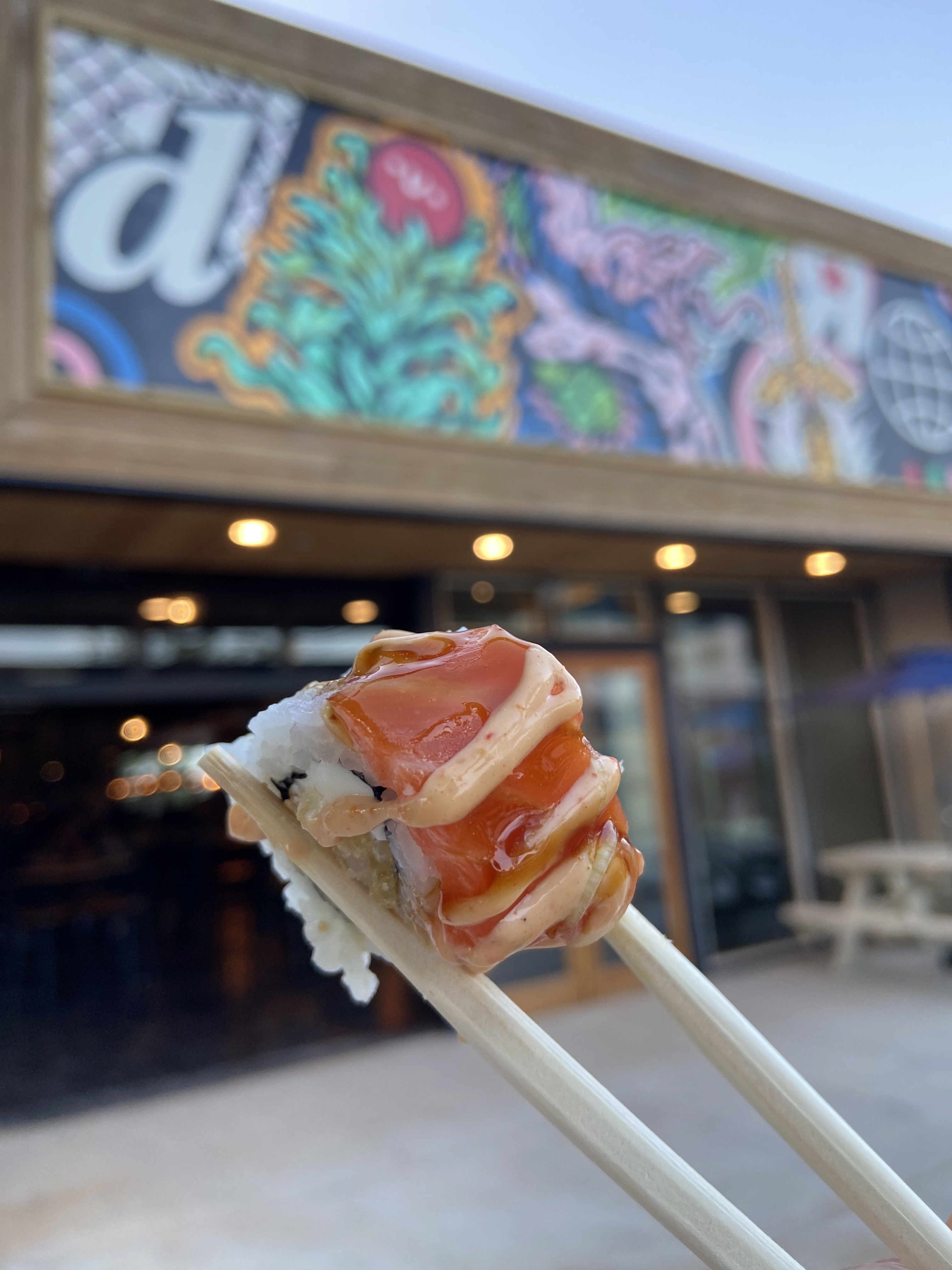 Close-up of sushi with salmon and spicy mayo sauce held by wooden chopsticks in front of a colorful mural and a restaurant exterior with lights.