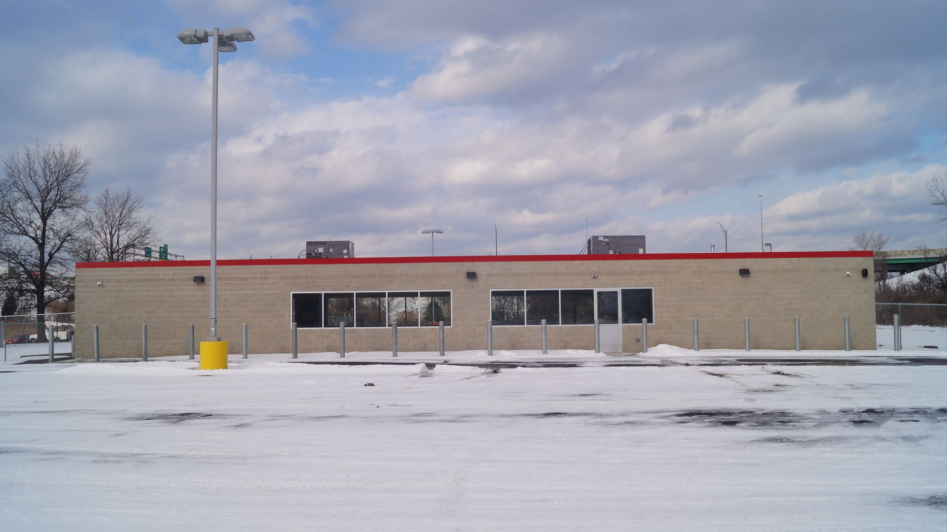 Snow-covered parking lot in front of a one-story beige brick building with a red roof trim, windows, and metal posts. Cloudy sky with patches of blue overhead.
