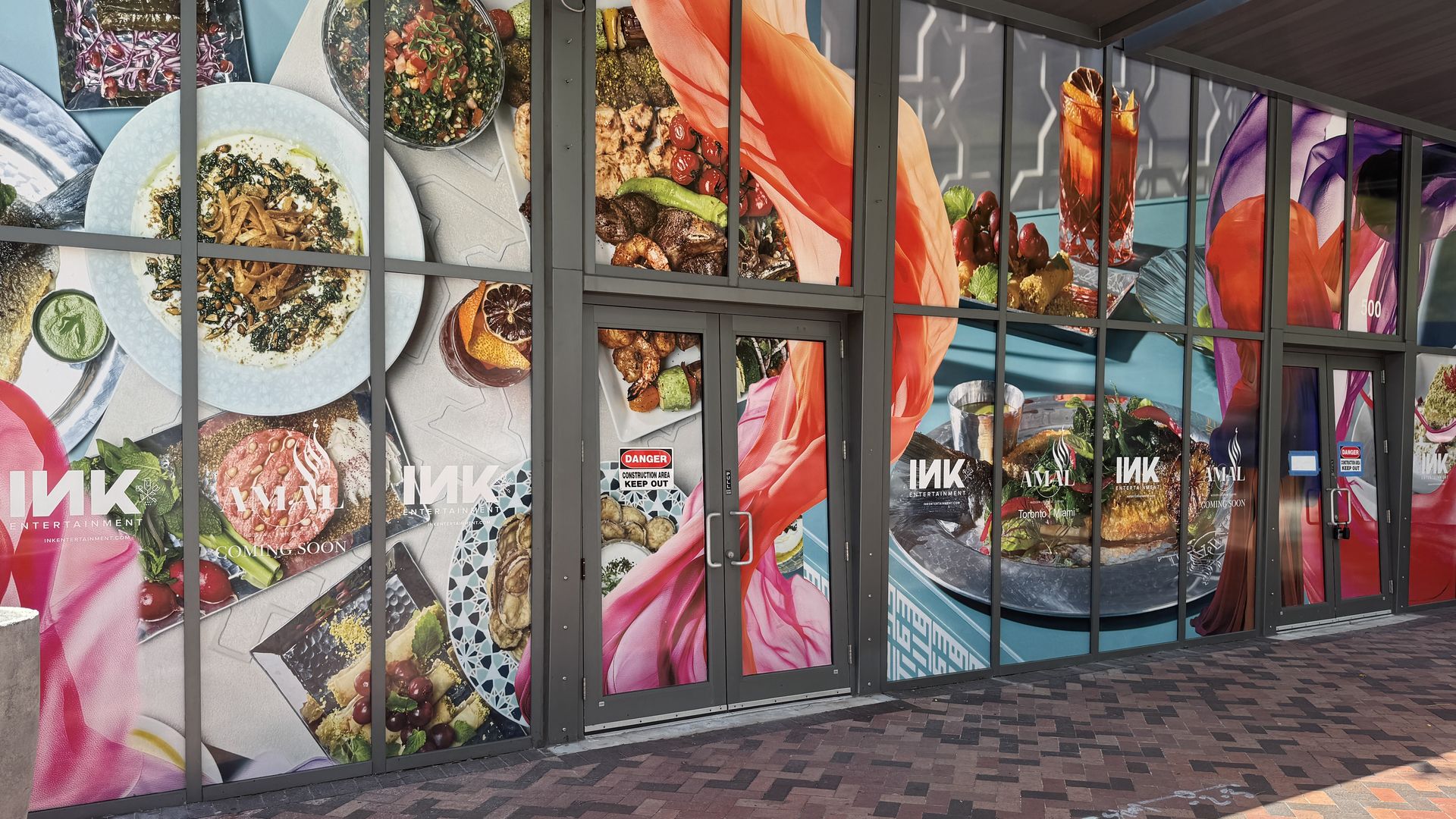 Colorful storefront wall on las Olas covered with large food photos—bowls, plates, and drinks—behind gray window frames; and a brick sidewalk lies in front.