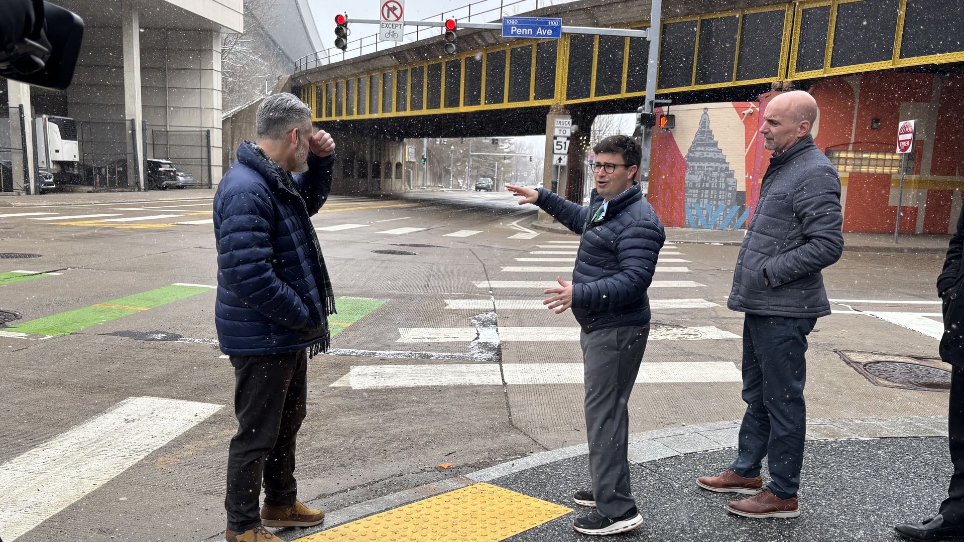 Three men in winter jackets on a snow-dusted city crosswalk under a yellow-railed overpass; one gesturing to another as the third looks on, with a mural and traffic signs nearby.