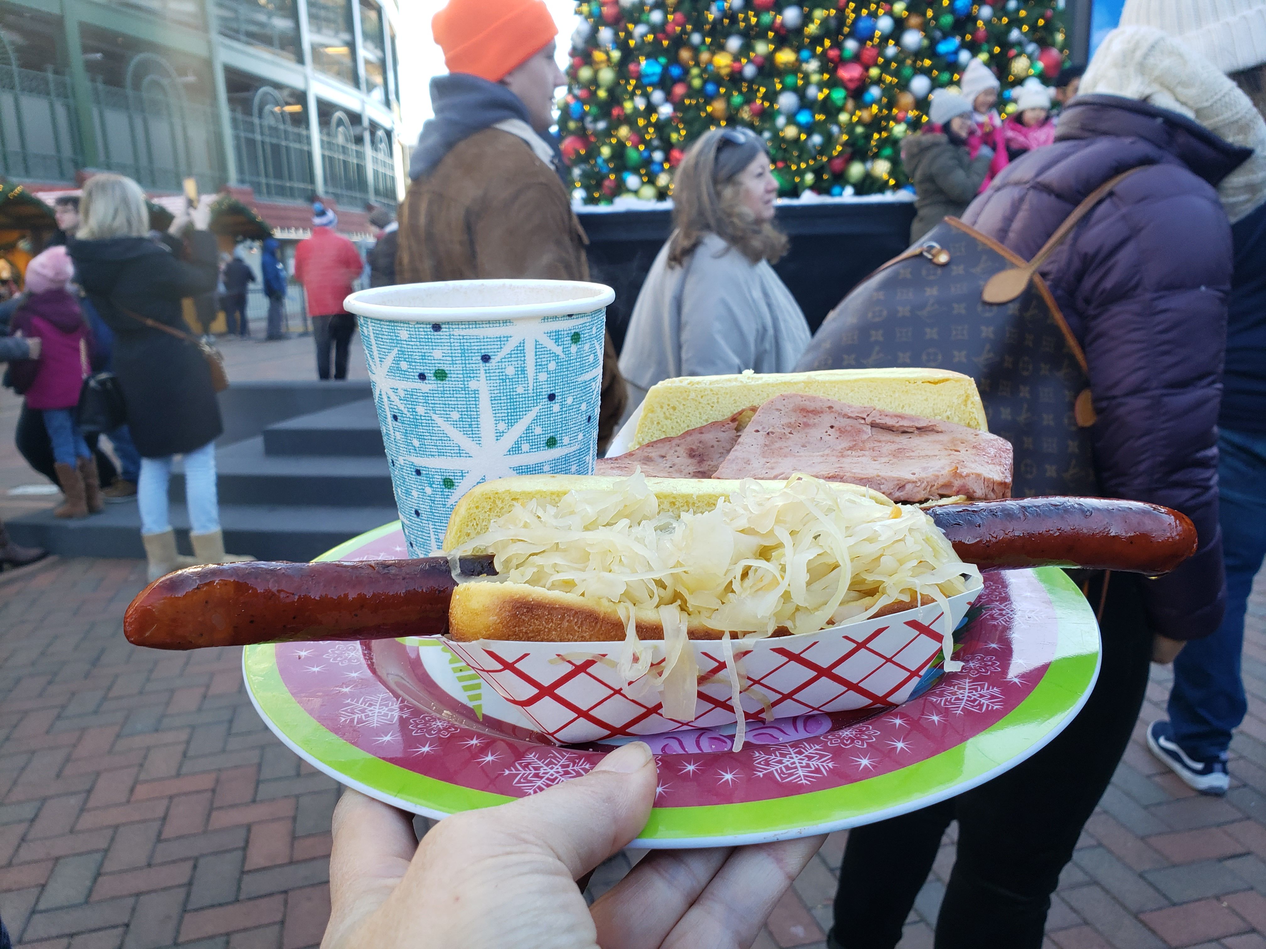 Hand holding a festive plate with a long sausage sandwich topped with sauerkraut, a sandwich with ham, and a blue cup with snowflake patterns in a crowded outdoor holiday setting with a decorated Christmas tree.