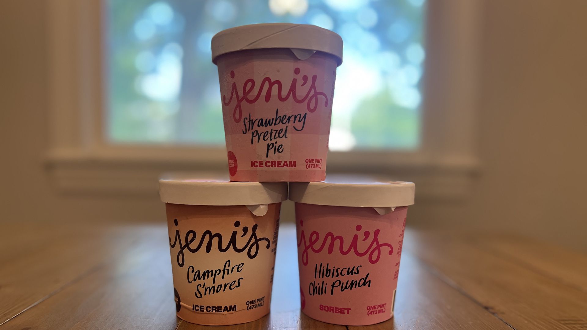 Four Jeni's ice cream cups stacked on a wooden floor with pink labels and white lids. The top cup reads "strawberry pretzel pie"; other flavors shown are "Campfire S'mores" and "Hibiscus Chili Punch," with a blurred window in the background.