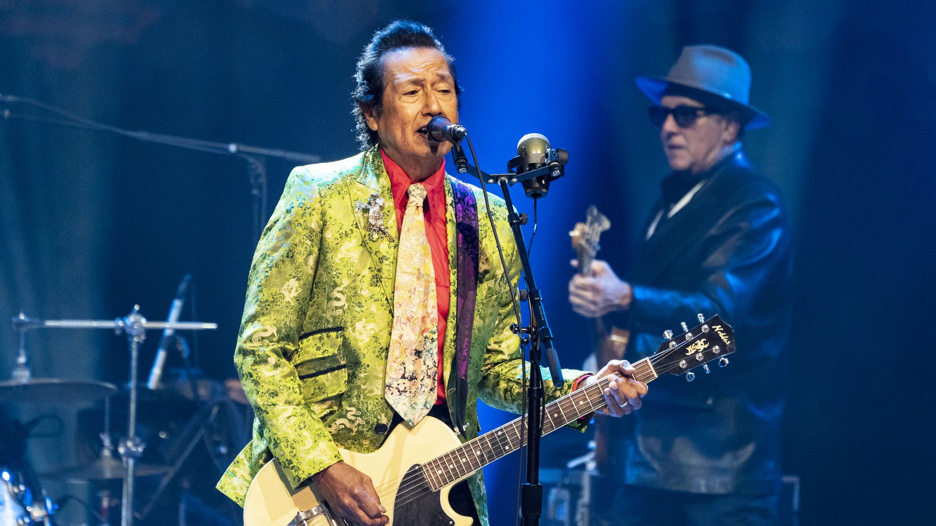 Male guitarist in a bright green patterned suit and red shirt sings into a mic on stage, playing a cream guitar; a second guitarist in a hat and sunglasses stands in blue light.