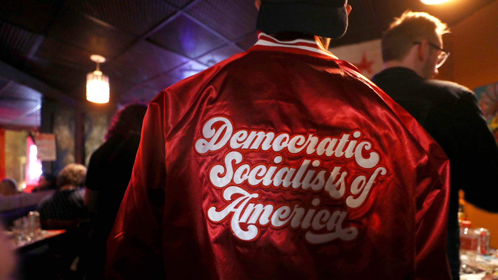 A view of the back of a red varsity-like jacket on a person, with the words "Democratic Socialists of America," in a bar setting