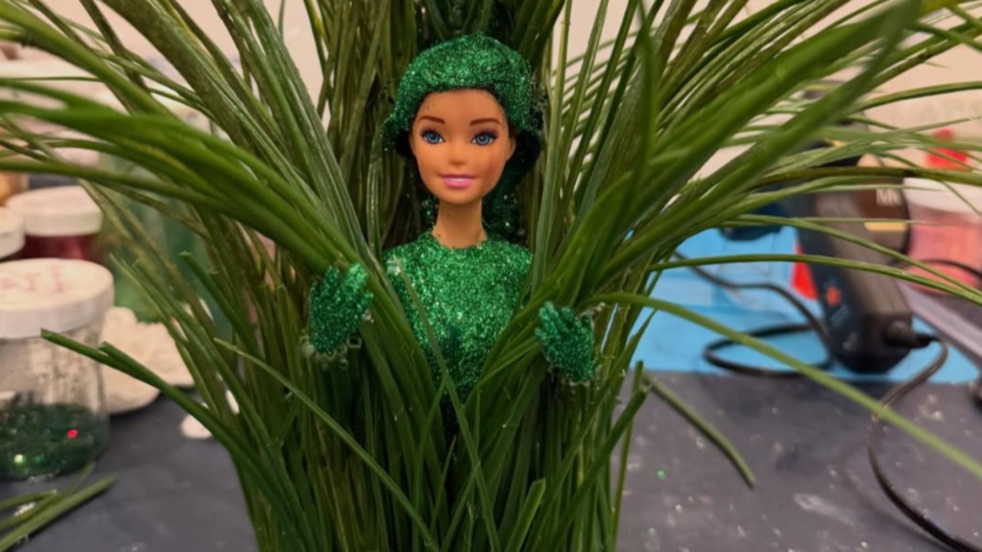 A Barbie doll covered in green glitter peeks through tall green plant leaves, holding some. Background shows jars and tools on a workspace table.
