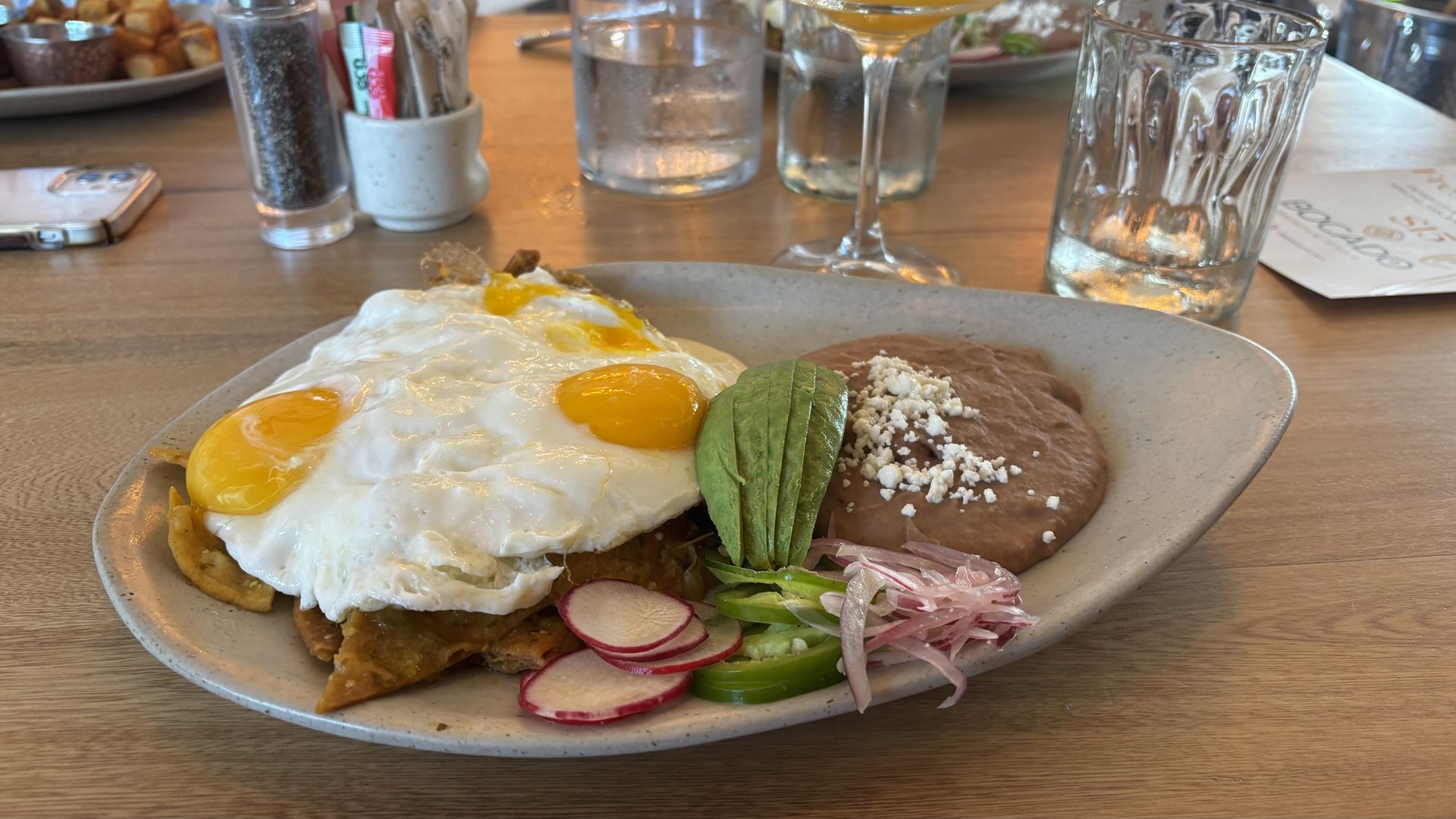 Plate of Mexican food with two sunny-side-up eggs, avocado slices, pickled onions, sliced radishes, jalapeños, and refried beans topped with cheese on a wooden table.