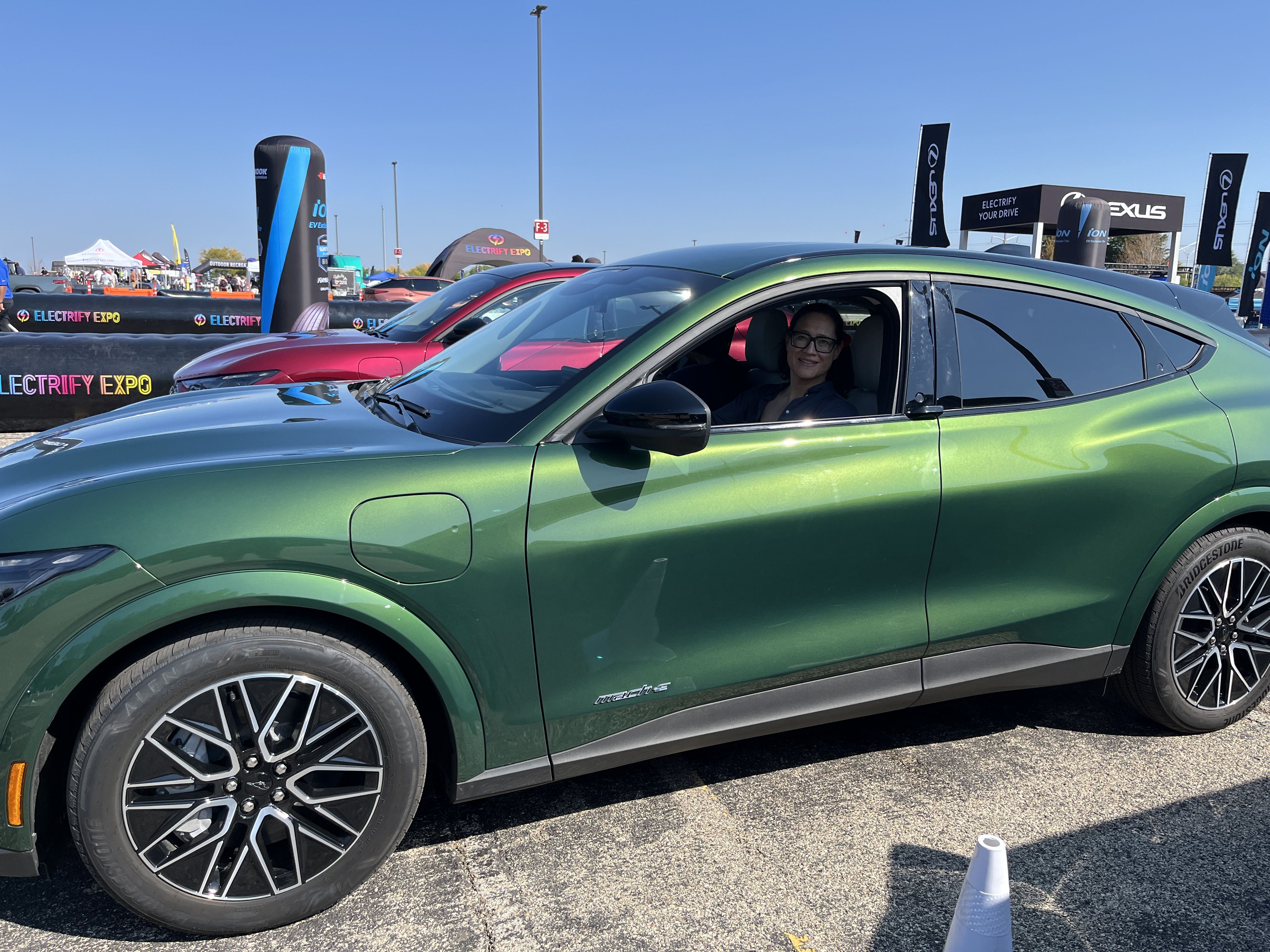 Green Mustang Mach-E electric SUV with a person smiling inside, parked at an outdoor Electrify Expo event with other cars and branded banners visible under a clear blue sky.