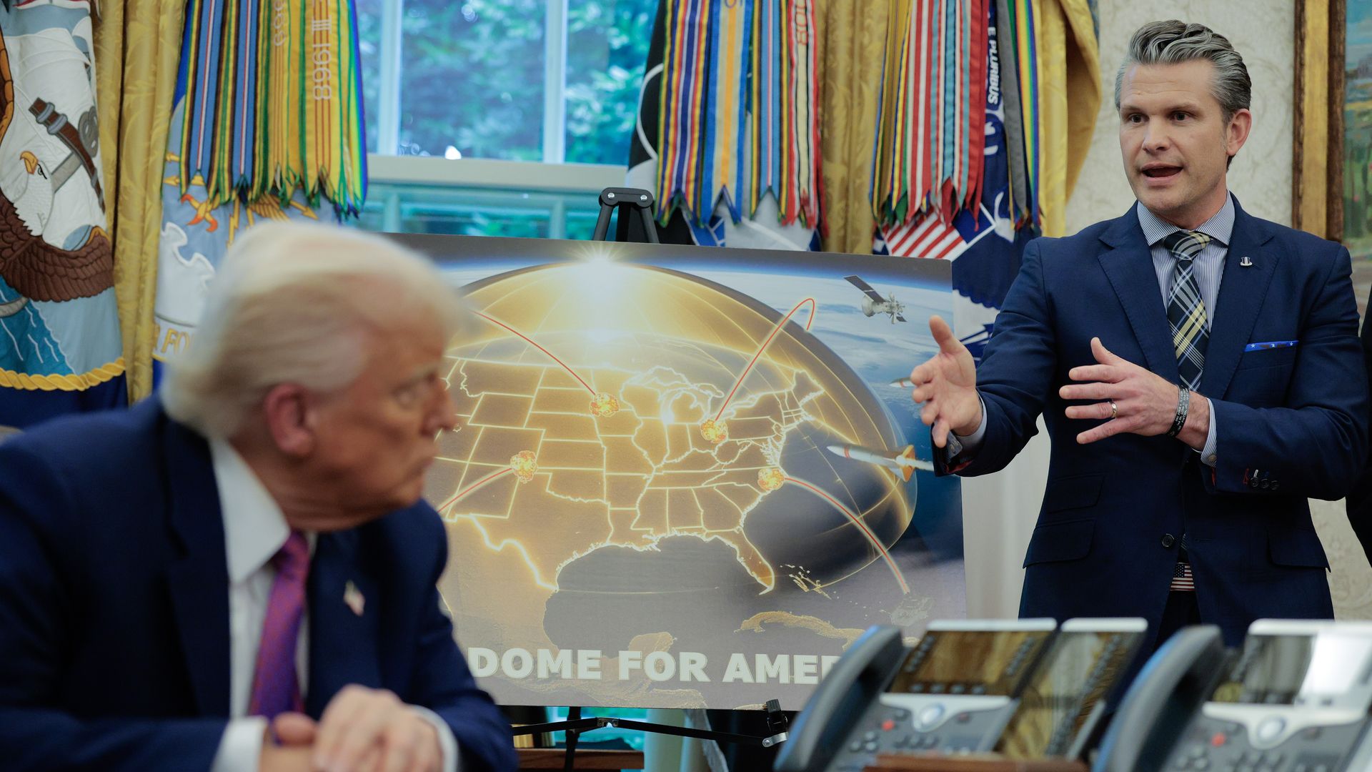 Pete Hefseth in a blue suit speaks and gestures to Donald Trump beside a glowing globe poster with route lines; colorful banners and flags fill the background.