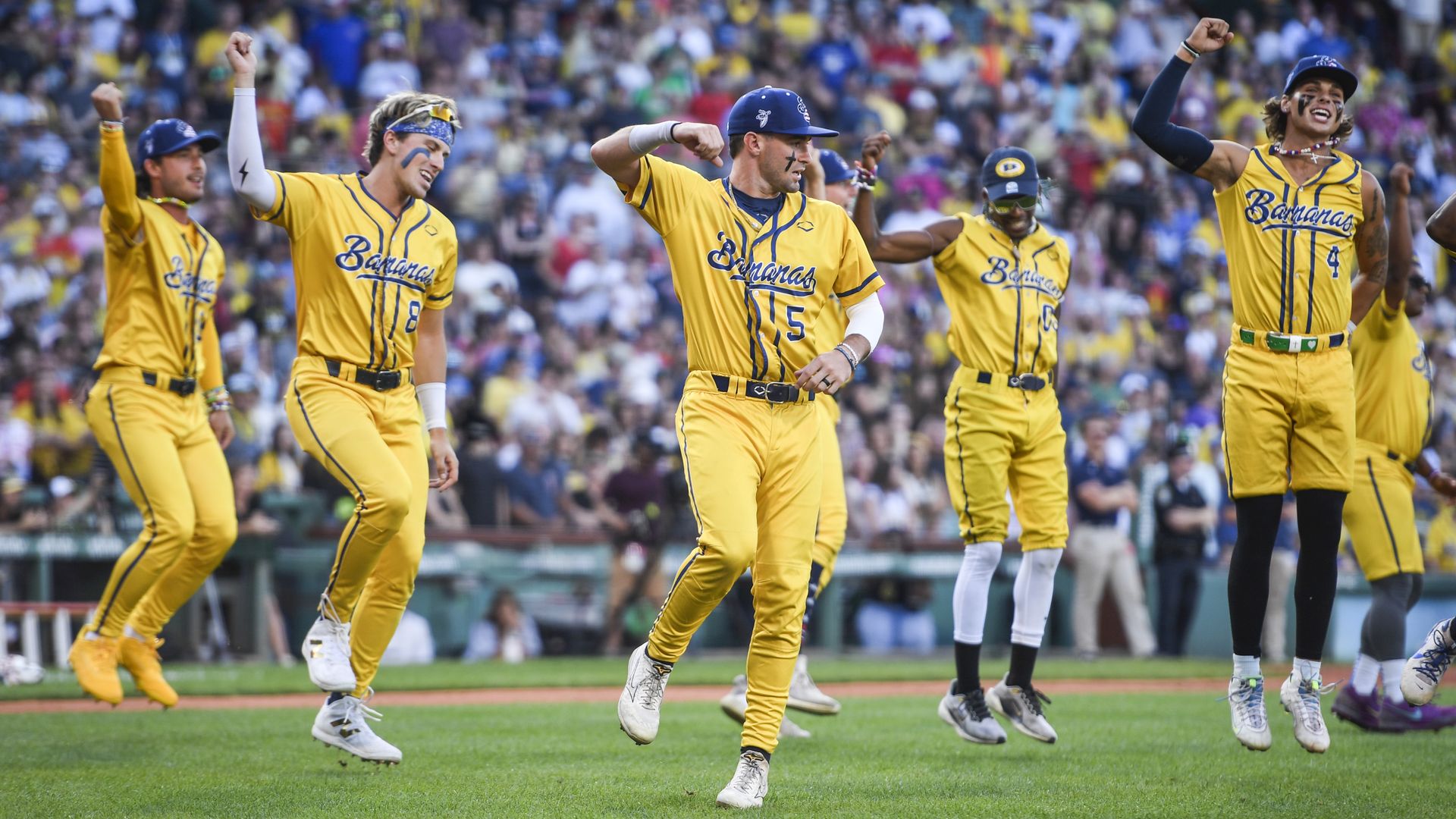 Five baseball players wearing blue caps and yellow uniforms with "BANANAS" on the front dance in unison on a baseball field, with a large crowd visible in the background.