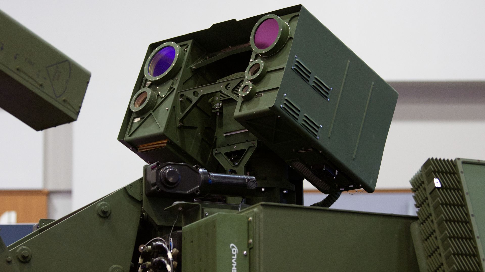 A laser weapon is mounted to a Stryker combat vehicle. It is an Army green color. The background is sparse, but mainly white.