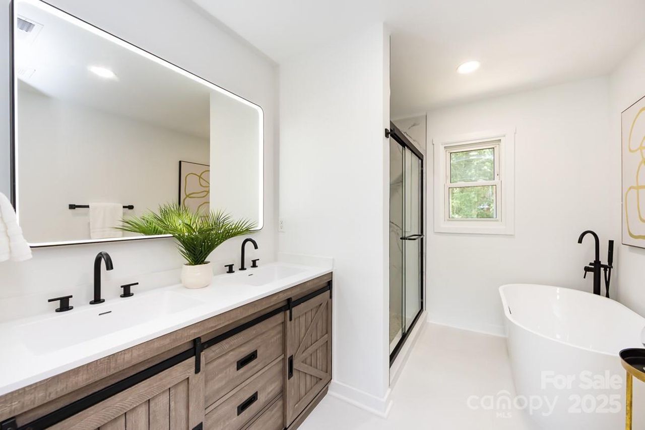 Modern bathroom with double vanity featuring wood cabinets, black faucets, a large illuminated mirror, a potted green plant, a glass shower enclosure, and a white freestanding bathtub with black fixtures.