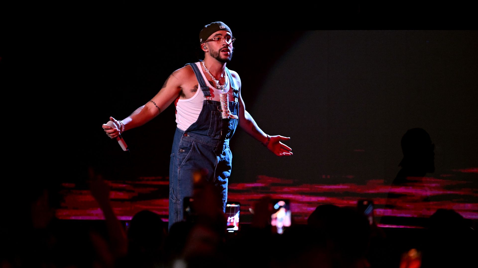 The musician Bad Bunny in denim overalls and a backwards baseball cap holding a mic.