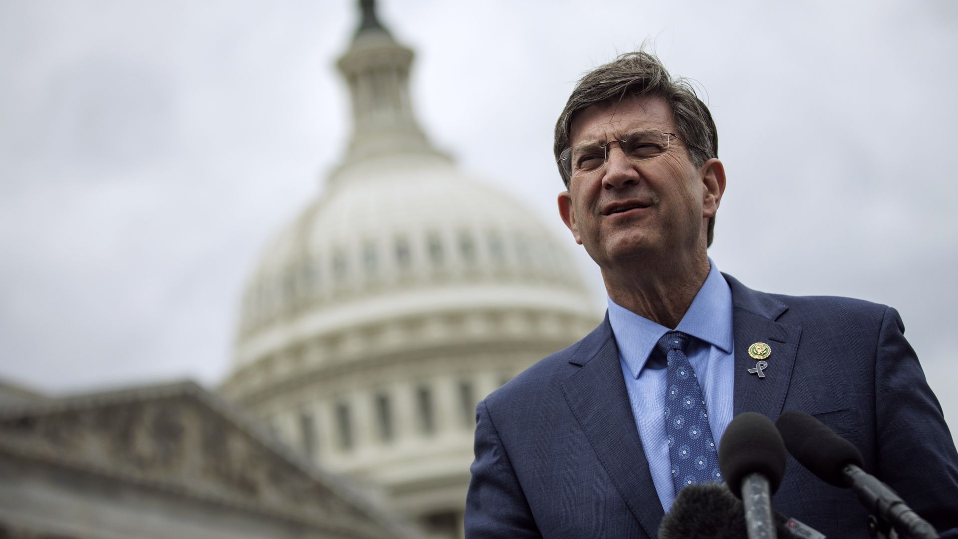 Rep. Brad Schneider, wearing a blue suit, blue shirt, blue tie and glasses, standing at microphones in front of the U.S. Capitol.