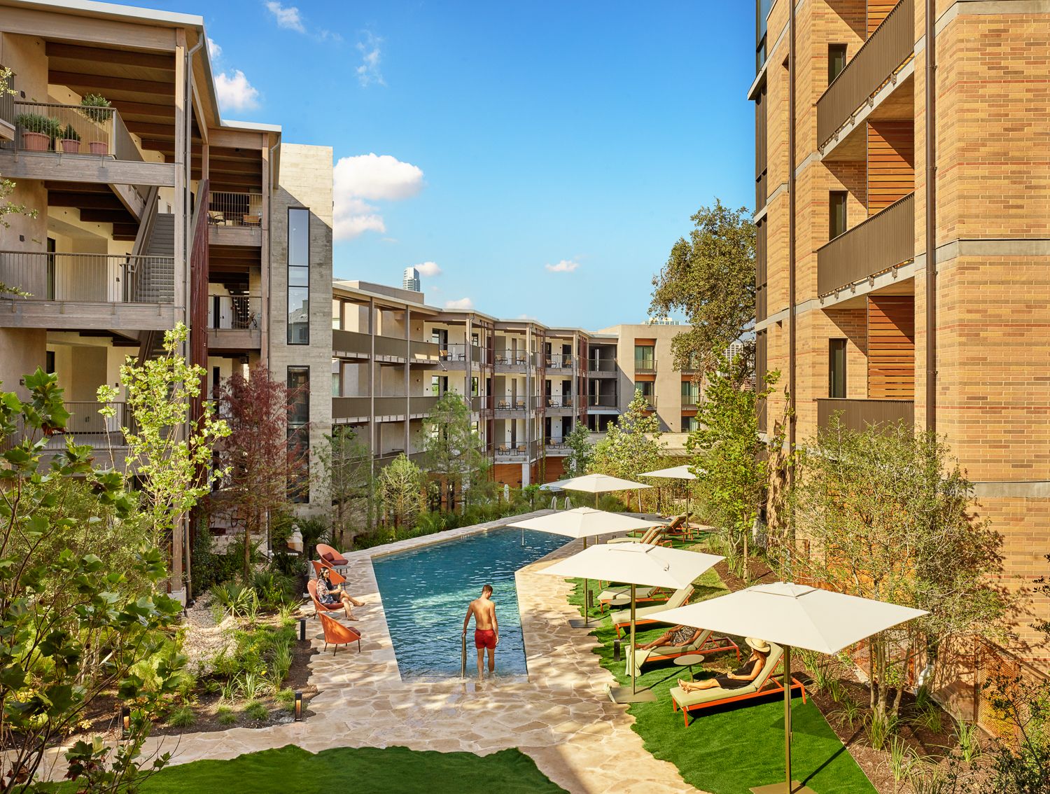 Sunny apartment complex courtyard with a rectangular pool, white umbrellas, and lounge chairs. Surrounding beige brick buildings and trees; a man in red trunks stands in the pool.