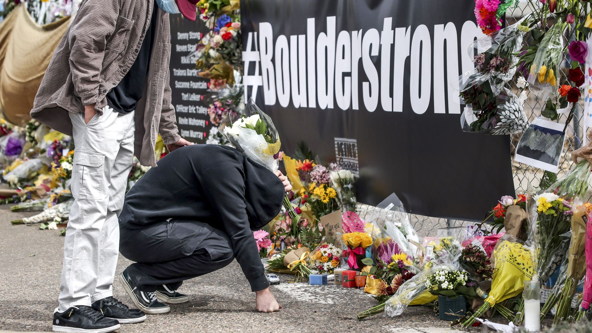 Photo of a person kneeling in front of a memorial with flowers and a sign that says "#BoulderStrong" as another person stands behind them with a hand on their back