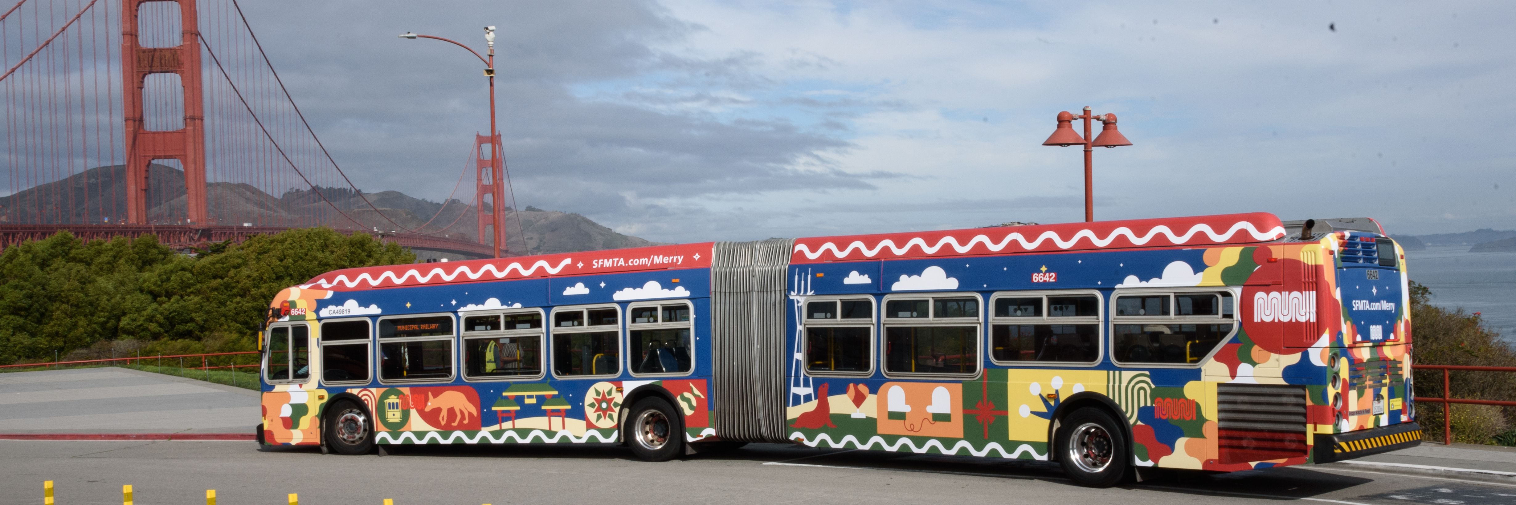Colorfully painted San Francisco Muni articulated bus with designs of landmarks and symbols parked near Golden Gate Bridge under cloudy sky.
