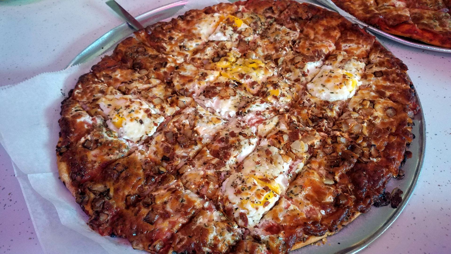 Large round pizza on a metal tray with a browned crust, topped with melted cheese, onions, herbs, and egg yolks; a second pizza is visible in the background.