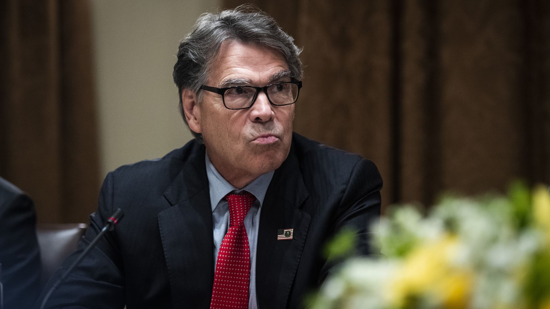 In this image, Rick Perry frowns and sits with his arms crossed while wearing a tie and glasses.