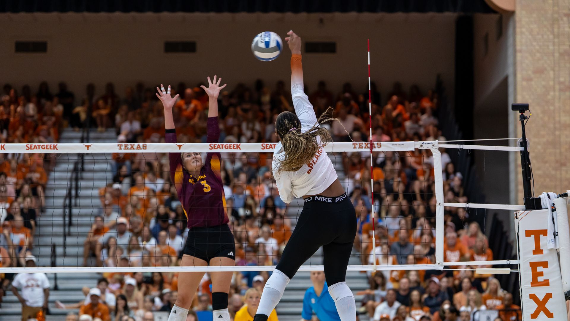 A Texas volleyball player in a white shirt spikes the ball as an Arizona State player tries to block it.