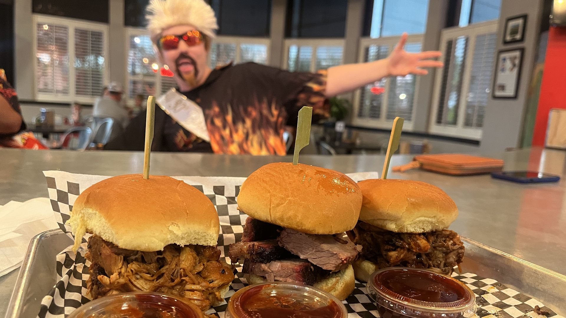 Three barbecue sliders with someone dressed like Guy Fieri in the background.