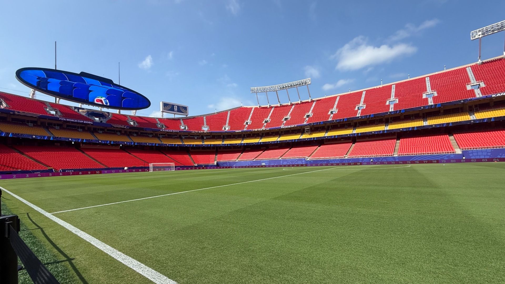 An empty soccer stadium with red seats and yellow sections, a large blue oval video screen hovering over the left stands, a green pitch, white goal in the distance, and a clear blue sky.