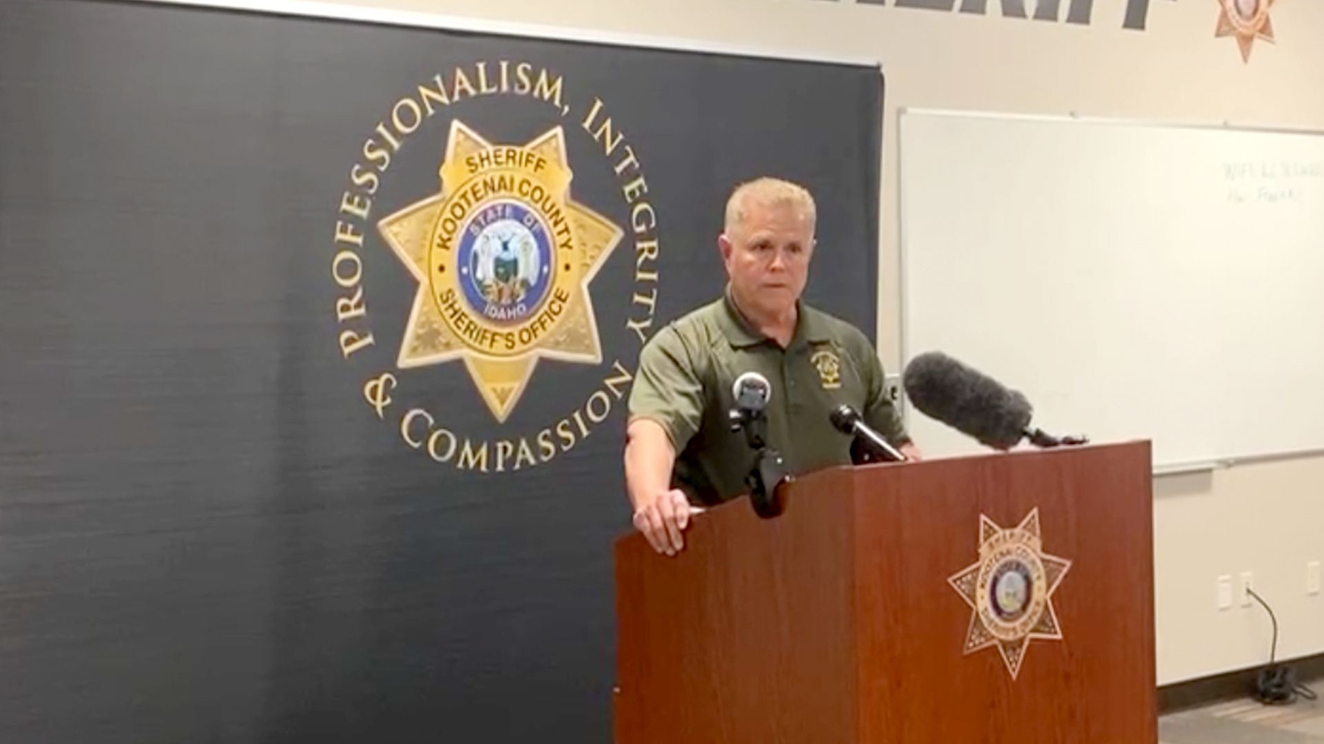 Kootenai County Sheriff Bob Norris speaking during a press briefing Monday afternoon, holding onto a wooden podium. The sheriff's office logo is behind him, featuring the words "professionalism, integrity & compassion" in gold writing, set against a black background.